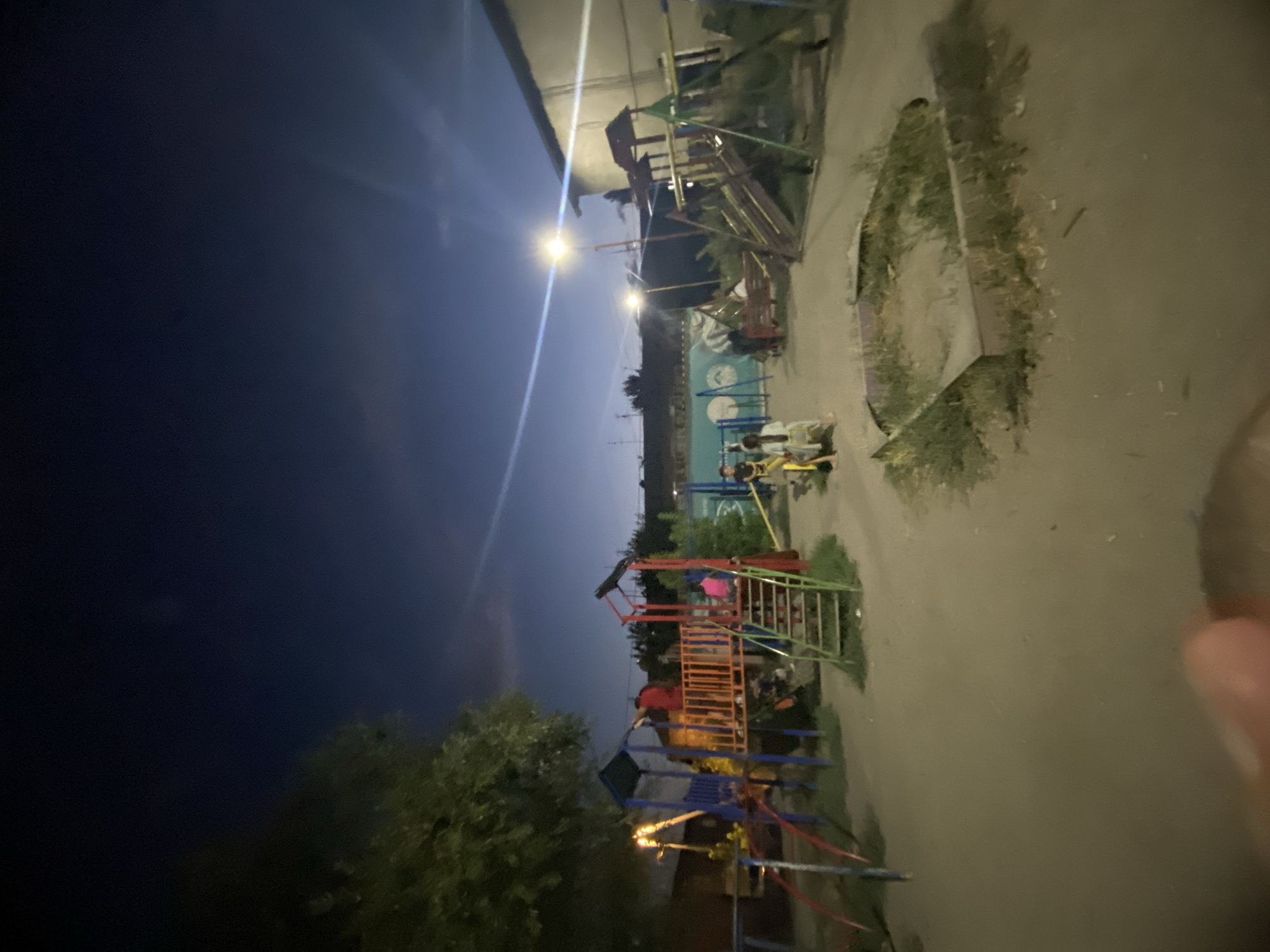 The image depicts a nighttime scene of an outdoor playground in Novopavlovka, Kyrgyzstan. The sky is dark blue with a hint of clouds. A few streetlights illuminate the area, casting long shadows. The playground features several structures, including a blue and red slide with yellow support beams, a red and orange platform with a green ladder, and a smaller yellow and blue see-saw. There are a few people visible: two individuals in the foreground on the see-saw, one wearing a light-colored outfit and the other a dark shirt. Another person in a red shirt is standing on the platform of the larger play structure, and a person in a pink shirt is seated on the platform. A few other figures are dimly visible near other play equipment. In the background, there are buildings and trees. The ground is mostly dirt and sand. The overall impression is of a community gathering space at night.