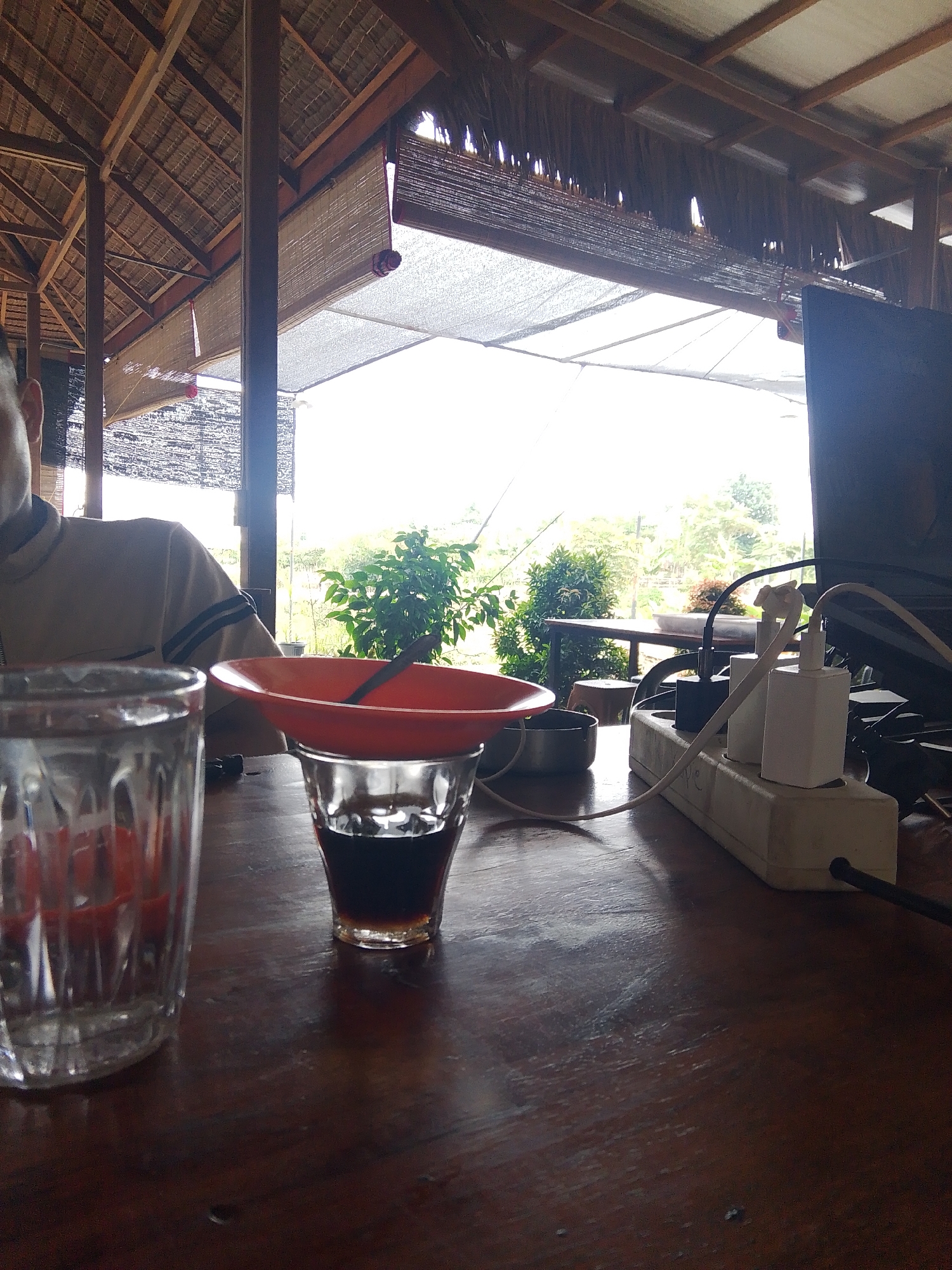 The image depicts an outdoor seating area, possibly a cafe or a warung, with a thatched roof supported by wooden beams. Bamboo blinds hang from the roof structure, partially obscuring a bright, overcast sky. A dark wooden table occupies the foreground. On the table, from left to right, are two drinking glasses: the left one is empty and ribbed, with a red object visible inside; the right one contains a dark liquid, possibly coffee or tea, and has a small orange bowl with a spoon resting on its rim. Behind the glasses, to the right, is a power strip with several white and black electrical adapters and cables plugged in. A black monitor is partially visible in the far right of the frame. In the mid-ground, a person wearing a collared shirt is partially visible from the shoulders up. The background shows lush green vegetation and trees, suggesting a rural or semi-rural setting. The overall impression is of a casual, open-air space. The provided location, Meunasahmee, Indonesia, places this scene in a specific geographical context.