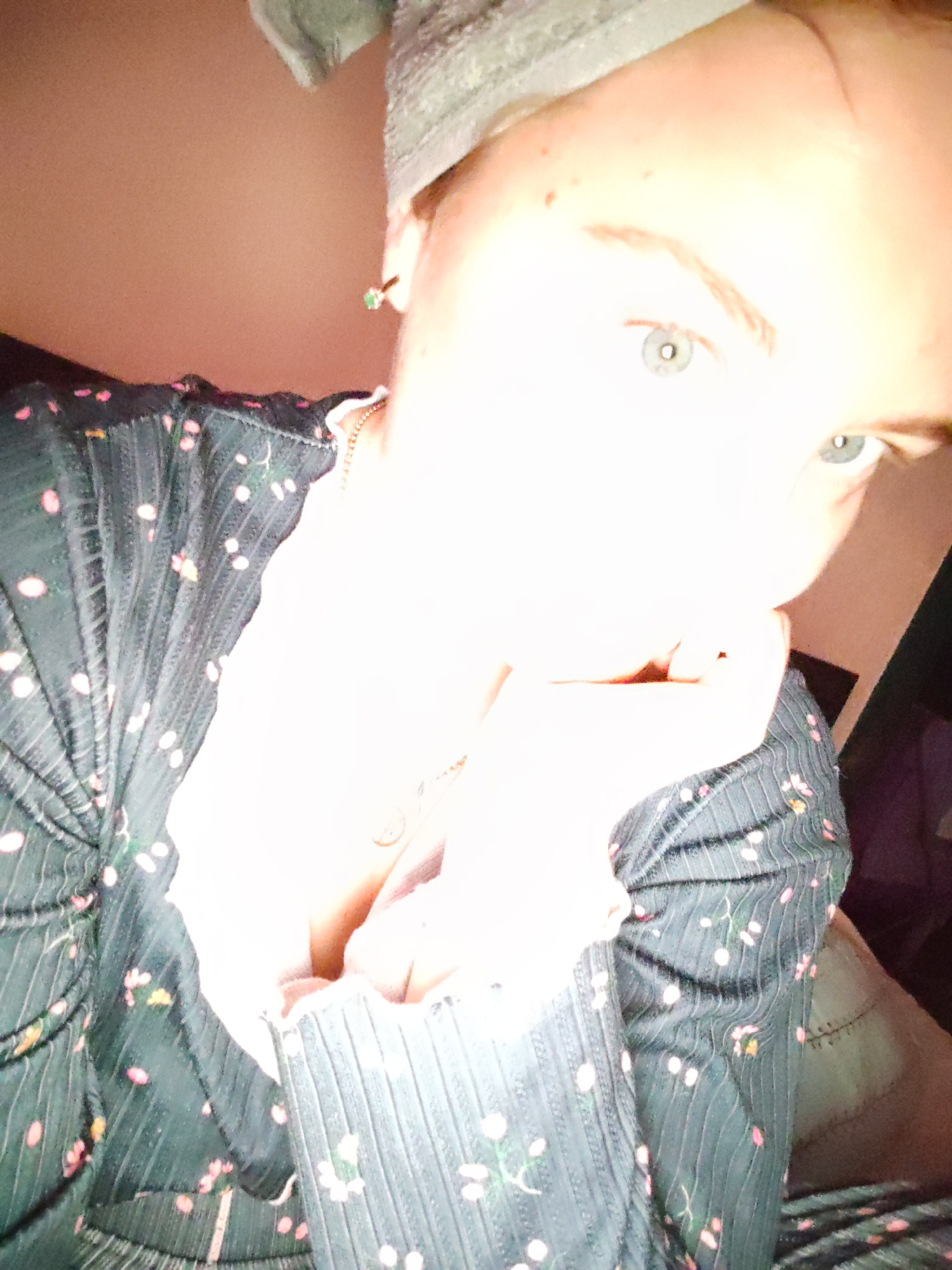 A person with blue eyes is facing the camera. The person is wearing a dark-colored garment with a small floral pattern. They are wearing a cloth head covering and an earring with a green stone. A necklace is visible. The background is blurry. It is not possible to determine the location as Izhevsk, Russia, without further information.