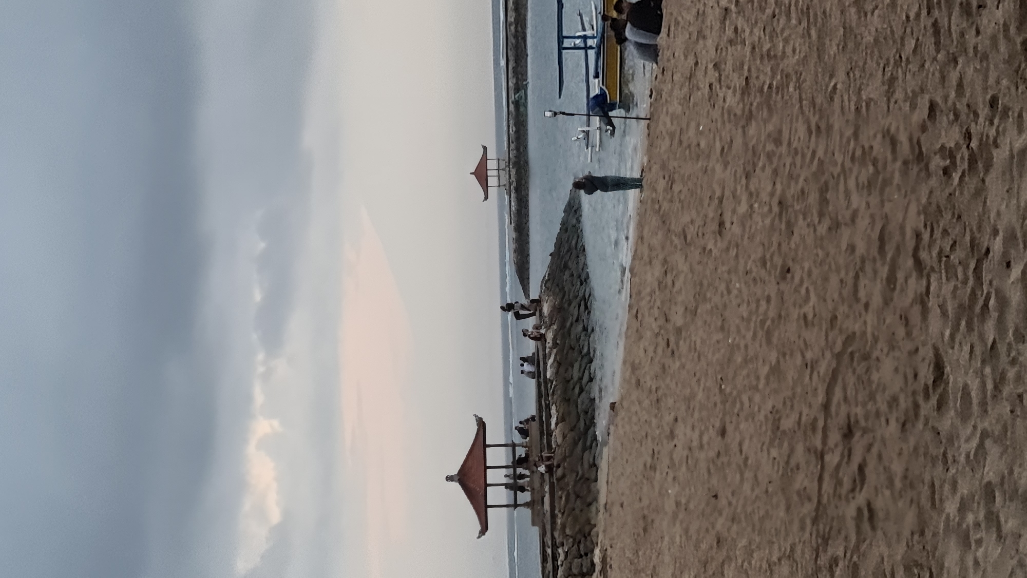 The image depicts a beach scene in Denpasar, Indonesia, likely at Sanur Beach given the presence of characteristic Balinese gazebos and outrigger boats. The sky is overcast with a soft, diffused light, suggesting either early morning or late afternoon. In the foreground, a sandy beach stretches out, with some footprints and undulations indicating the texture of the sand.
Further back, along a rocky breakwater, several people are seated and standing, some appear to be observing the water or conversing. A prominent red-roofed Balinese gazebo, supported by wooden posts, stands on this breakwater, offering shade. Beyond this, another similar, smaller gazebo is visible in the distance on a separate section of the breakwater.
The water is relatively calm, with gentle waves lapping at the shore and the breakwater. Several boats are moored near the beach. One is a blue and yellow outrigger boat with a covered seating area, and another appears to be a smaller white vessel. A person is standing on the wet sand near the water's edge, facing away from the camera, possibly taking a photograph. In the far right of the image, a group of people are seated on the sand, facing the water. The overall atmosphere is relaxed and serene.