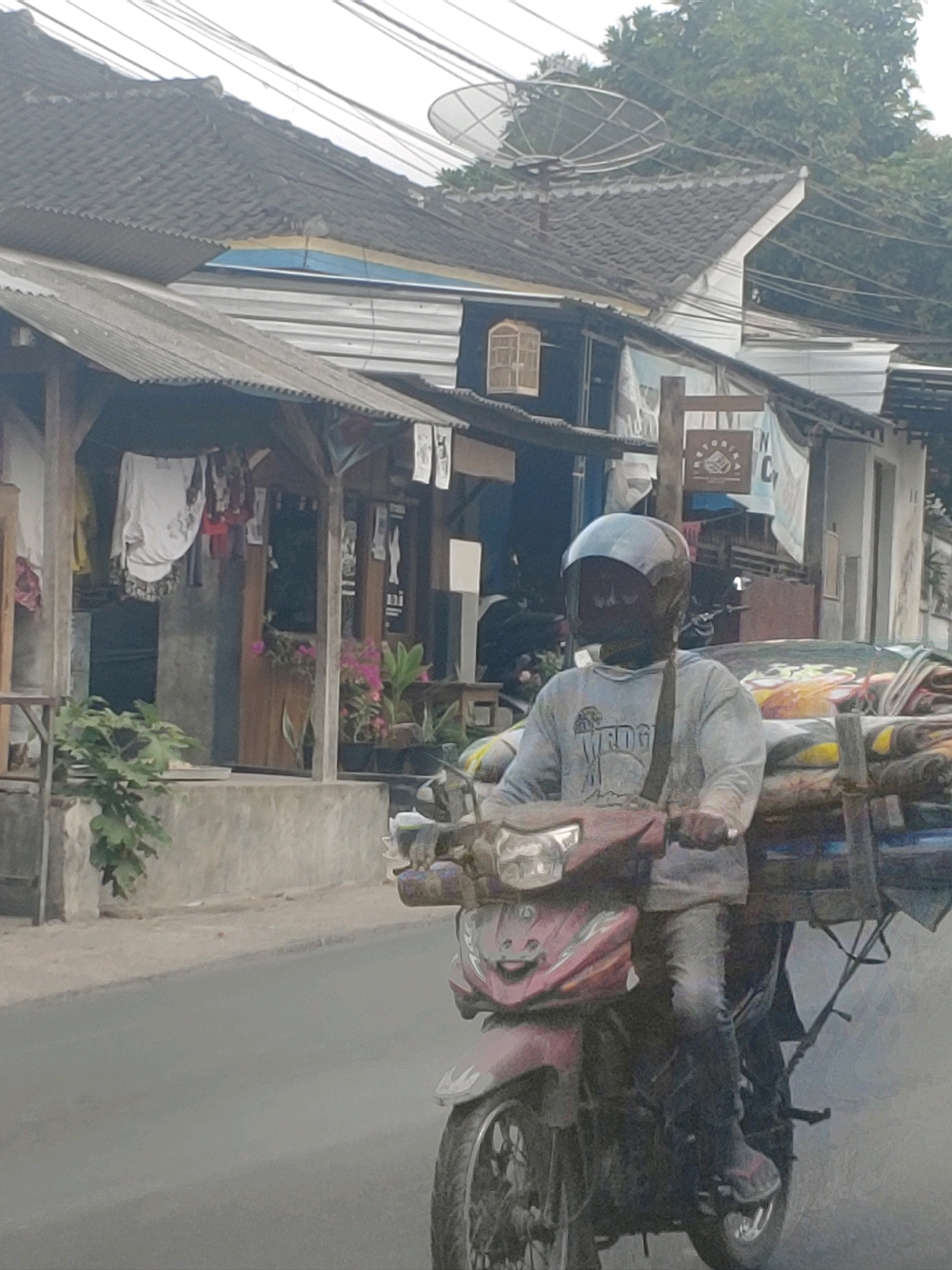 A man wearing a blue helmet and a light blue long-sleeved shirt rides a red motorcycle down a paved road in Kembang Kerang, Indonesia. The motorcycle has a large wooden rack behind the seat, loaded with what appear to be sacks of goods. The man is wearing dark pants and dark shoes. In the background, there are buildings with corrugated metal roofs and what looks like a satellite dish on one of the roofs. There are also power lines visible above the buildings and trees. The overall scene depicts a typical street in a populated area of Indonesia.