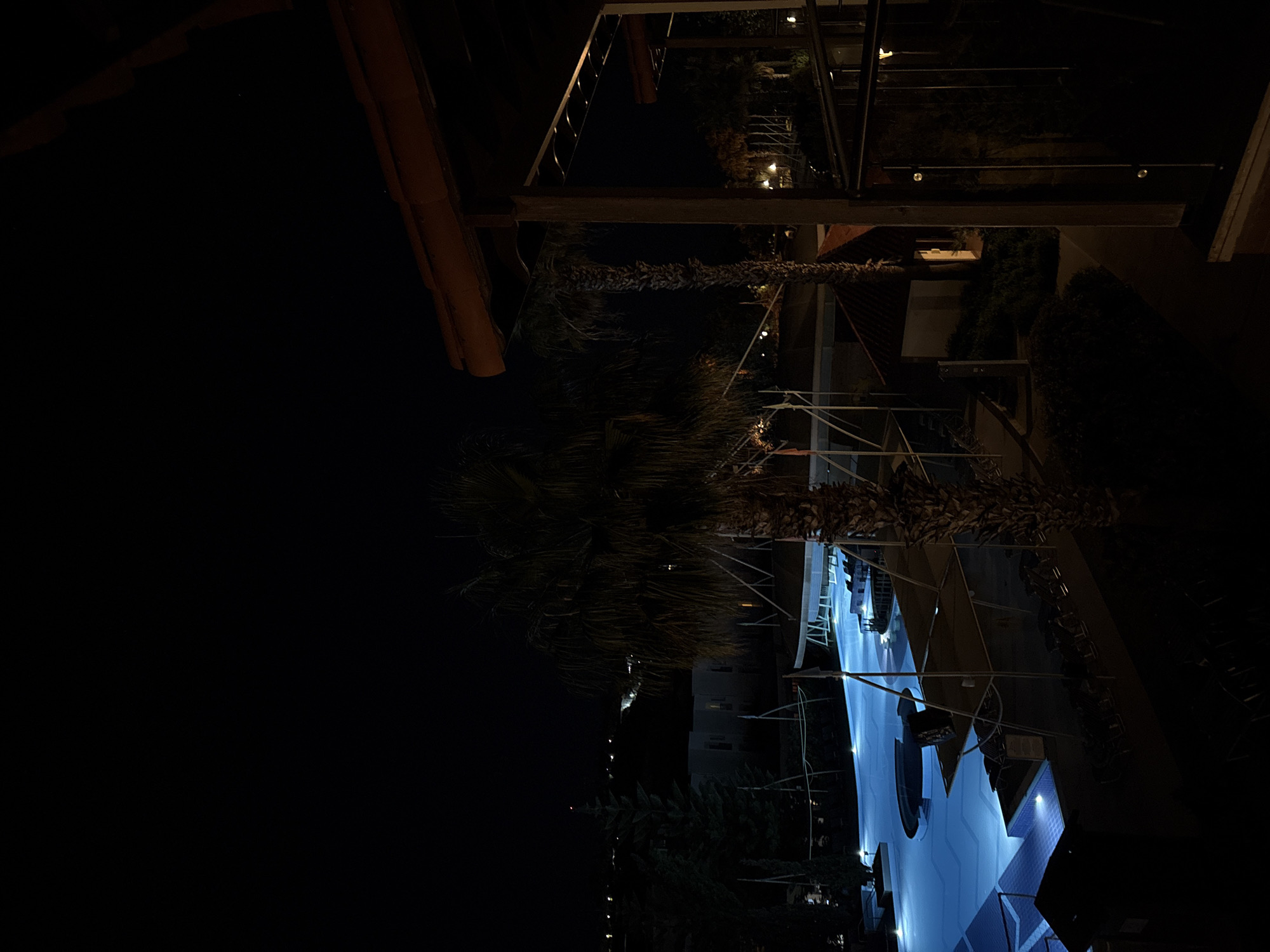 The image presents a nighttime view of an outdoor resort or hotel area, primarily centered around a large, illuminated swimming pool. The pool, characterized by its irregular shape and distinct blue lighting, is the brightest feature in the dark environment. Around the pool, numerous white lounge chairs and umbrellas are neatly arranged, indicating a leisure or relaxation zone, though no people are present.
Several dark palm trees and other dense green foliage are visible throughout the scene, their silhouettes prominent against the ambient lights. Architectural elements, possibly structures for overhead shading or balconies of nearby buildings, frame parts of the view, with some featuring warm-toned lights. In the background, dimly lit buildings with a few visible windows suggest accommodation or additional resort facilities.
The time of day is clearly night, with the sky being completely dark and all visibility relying on artificial lighting, which ranges from the bright blue of the pool to warmer ambient glows. The weather appears clear, with no visible clouds or precipitation. The overall atmosphere conveyed is one of tranquility and serenity, suggesting a peaceful evening at a holiday destination. No visible text is discernible in the image.