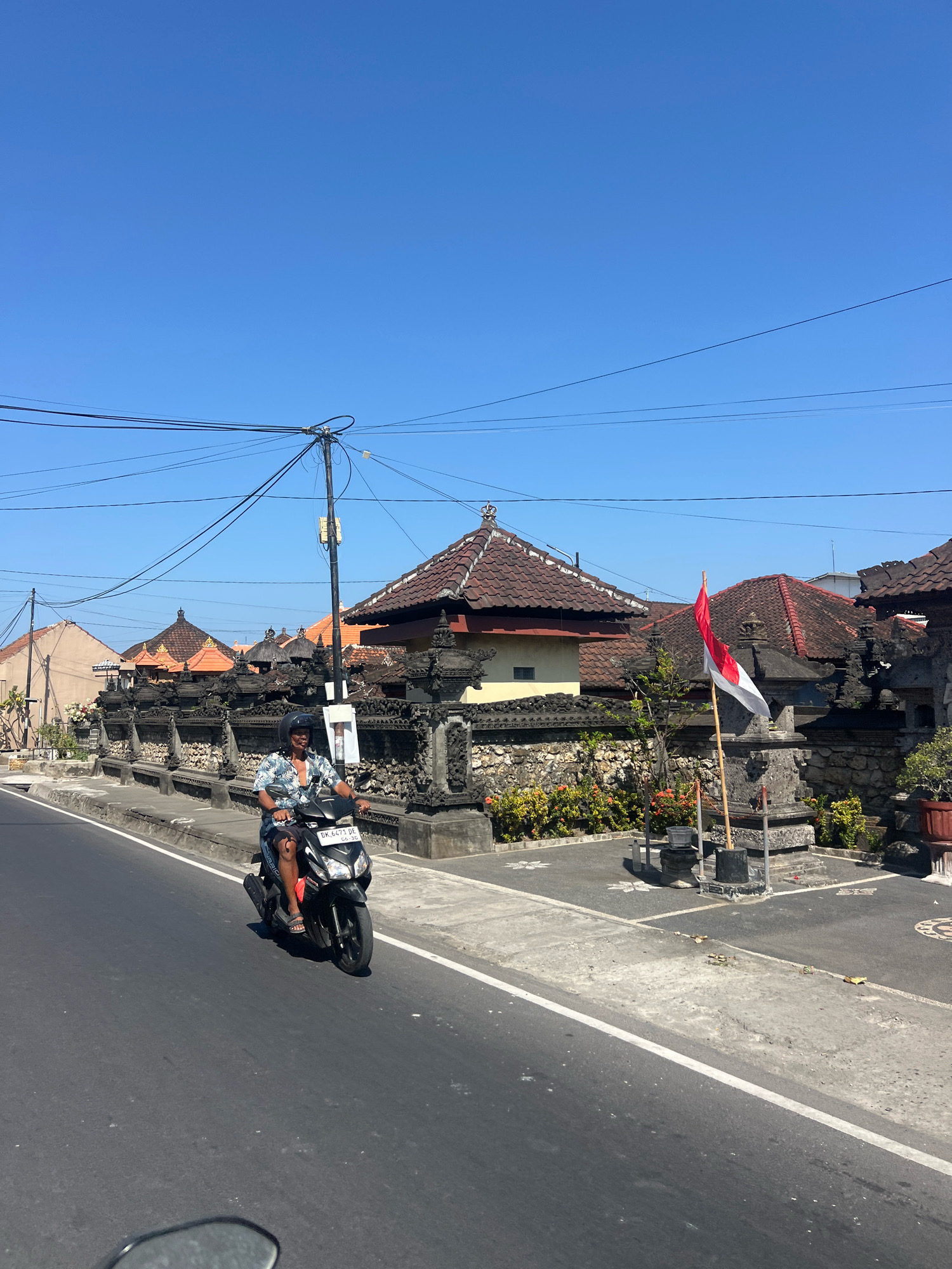 A man rides a scooter down a street in Denpasar, Indonesia, with a clear blue sky above. He is wearing a patterned shirt and shorts, with a helmet on his head. The scooter has a license plate that reads "DK 6473 DE".
The street is lined with a stone wall adorned with intricate carvings, typical of Balinese architecture. Behind the wall, traditional houses with tiled roofs are visible. A Indonesian flag, red and white, stands planted on a pole, fluttering in the breeze. Lush green foliage and flowering plants are seen along the roadside and within the property behind the wall. Power lines crisscross the sky, supported by utility poles. The road surface is asphalt, with a white line marking the lane. The scene appears to be a sunny day in a residential or temple area of Denpasar.