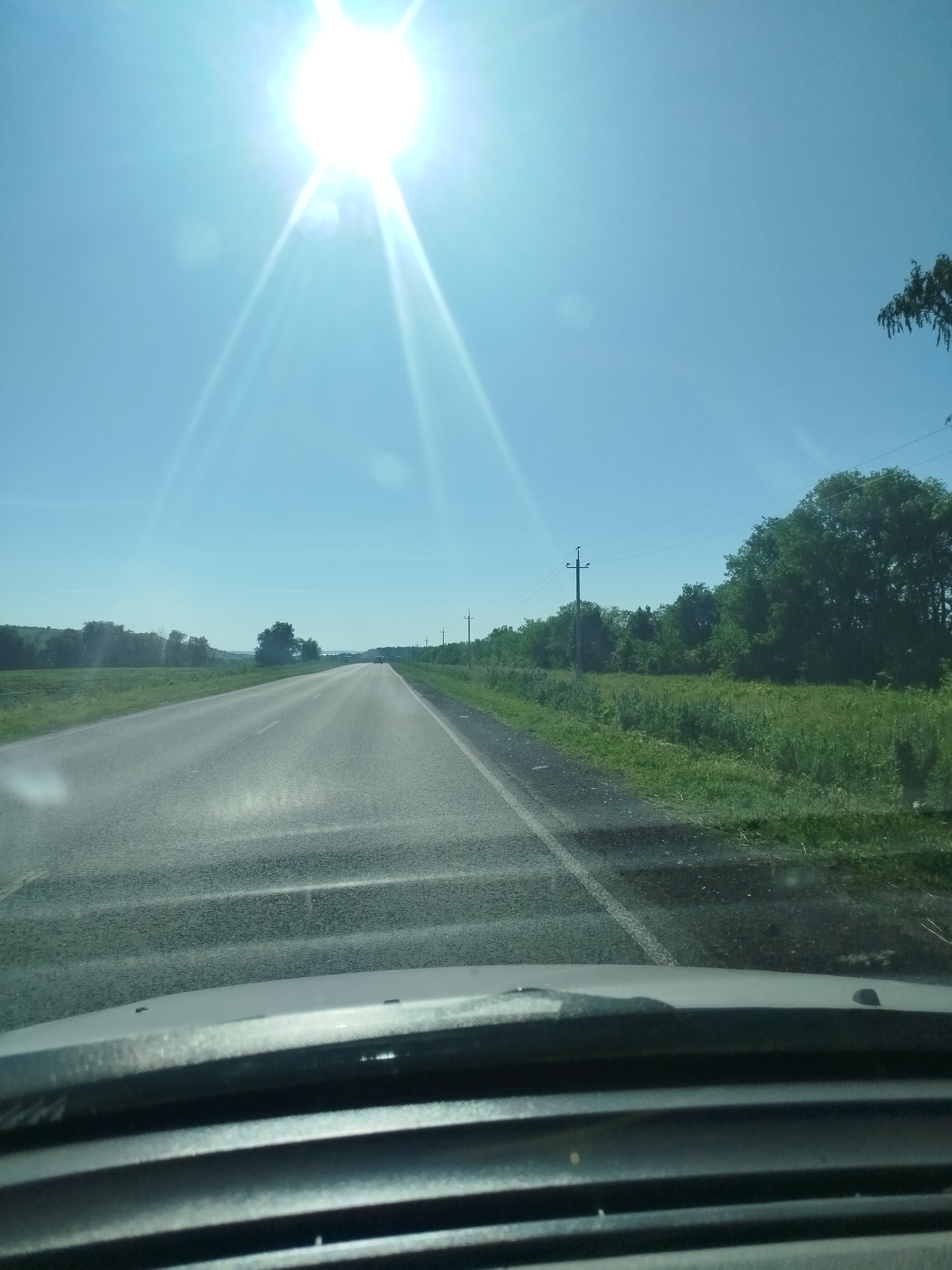 The image shows a view from inside a vehicle driving on a paved road under a bright, clear blue sky. The sun is high and appears to be slightly to the left of the frame, causing lens flare and bright streaks of light. The road is asphalt with a white line marking the right edge. On either side of the road, there are grassy fields and trees. Power lines and utility poles are visible along the right side of the road. In the distance, the road stretches towards the horizon, with a slight curve. The foreground shows the interior of the vehicle, including the dashboard and windshield. The overall impression is of a sunny day drive through a rural or semi-rural landscape. No specific details about the city of Tushna, Russia are visible in the image.