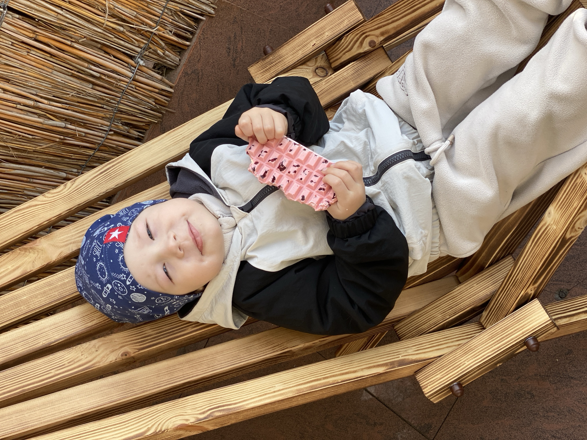 A young child wearing a blue patterned hat, a black and white jacket, and light gray pants lies on a wooden bench, holding a pink waffle-like treat. The background includes a reed-like structure and a textured surface, suggesting an outdoor setting in Sochi, Russia.