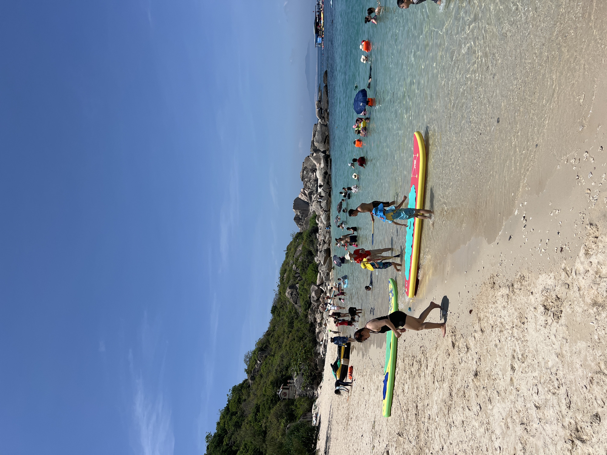The image depicts a crowded beach scene in Dai Lanh, Vietnam, under a bright, clear blue sky with minimal high-altitude clouds. The foreground features a wide expanse of light tan sand transitioning into remarkably clear, turquoise water. Numerous individuals, including adults and children, are present on the beach and in the water, engaging in recreational activities.
On the left, a steep, verdant hillside covered in dense green foliage and large grey boulders rises. A small, multi-tiered structure with a red roof, resembling a shrine or temple, is visible halfway up the hillside. Towards the center-left on the sand, a black and yellow inflatable boat or jet ski is beached, surrounded by a few people. Two brightly colored inflatable paddleboards, one green and one red and yellow, rest in the shallow water or on the wet sand in the lower portion of the image. A woman in a black swimsuit walks barefoot from the water towards the dry sand, positioned slightly left of center. Children are seen near the paddleboards, and one child in a blue shirt and patterned shorts stands in the water near the red and yellow board.
The water is teeming with people, most of whom are wading or standing in the shallow depths. Several individuals wear orange life vests. A person is seen holding a blue beach umbrella while standing in the water. In the background, large grey rock formations extend into the sea, providing a natural boundary to the beach. Beyond these rocks, the sea appears deeper blue, and several larger boats, likely tour vessels, are visible, some with blue canopies. In the far distance, faint outlines of hills or mountains are visible under a slight atmospheric haze. The scene is well-lit by direct sunlight, indicating a sunny day.