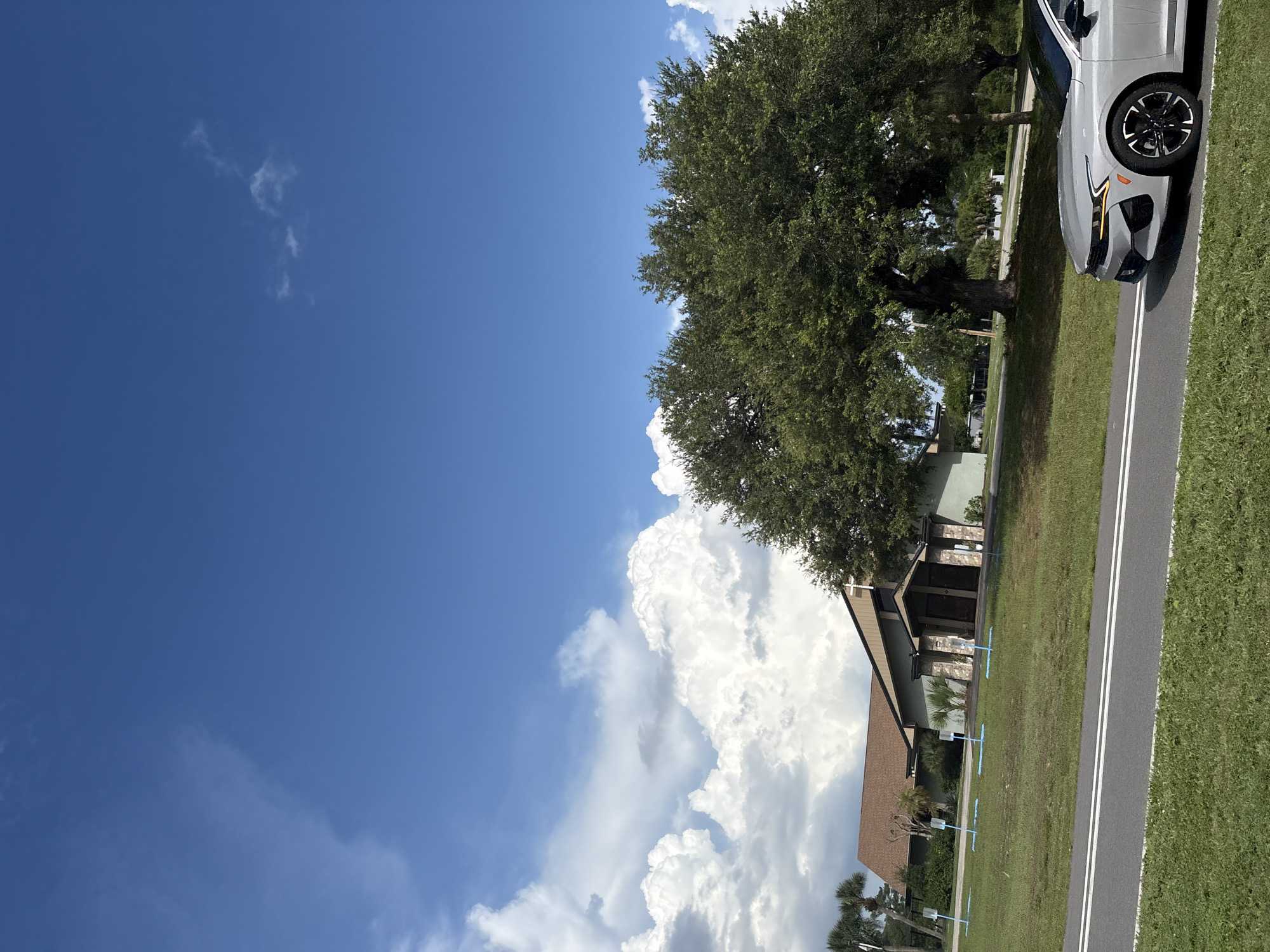 An exterior, ground-level, eye-level shot captures a scene in North Port, United States of America, on a bright, cloudy day. In the foreground, a modern silver car is parked on an asphalt road with white lane markings. Behind the road, a well-maintained grassy lawn slopes up towards a light-colored, single-story building with a dark-pitched roof. The building features a prominent entrance with two doors, flanked by stone pillars and a cross at the top. Several blue signs with white markings are visible on the lawn in front of the building, spaced evenly apart. To the right of the building, a large, mature tree with dense green foliage dominates the middle ground. The background is filled with a bright blue sky punctuated by large, fluffy white clouds, some of which are illuminated by the sun. Palm trees and other vegetation are visible on the left side of the building.