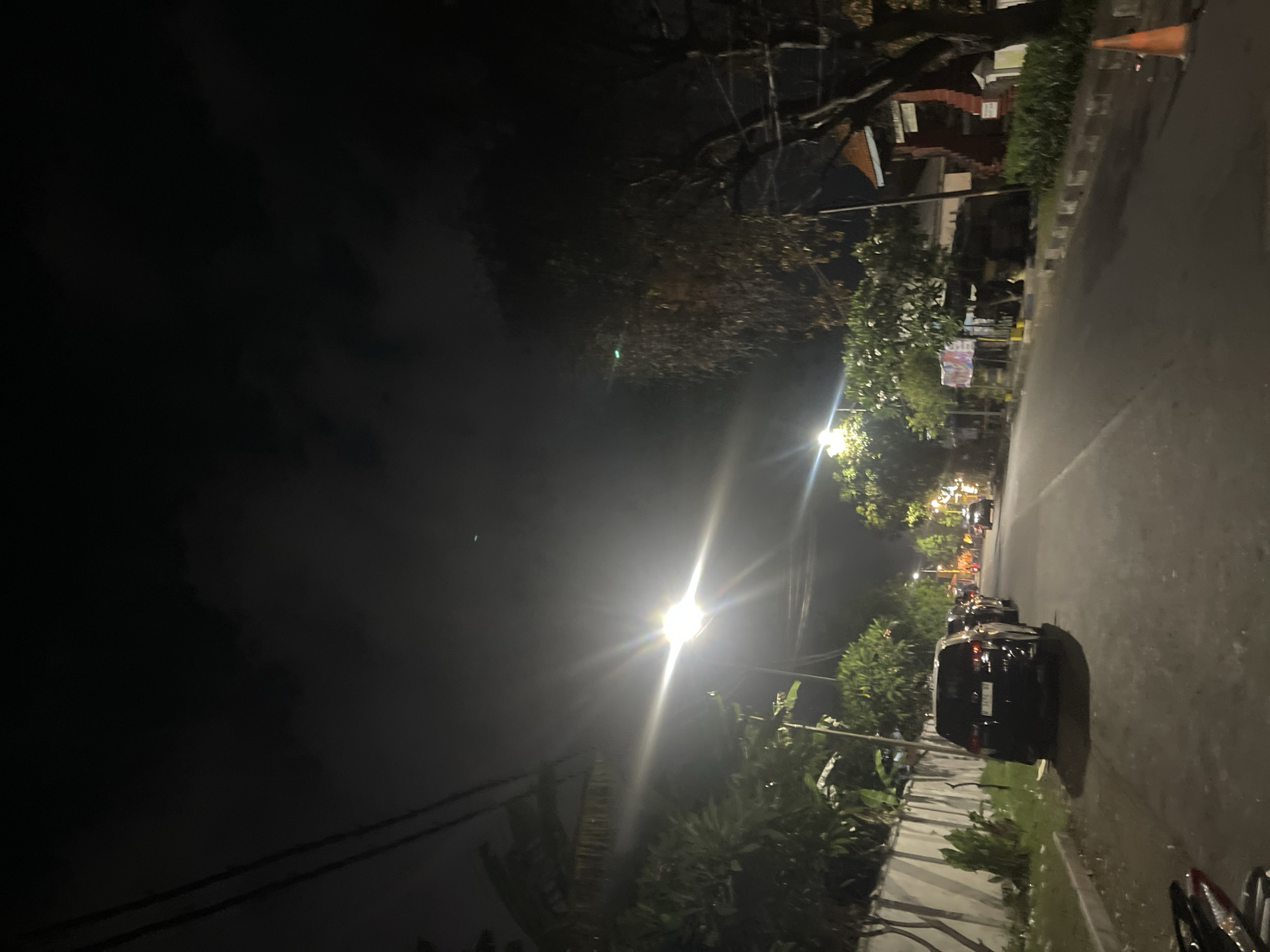 This image depicts a dimly lit street scene at night in Sidoarjo, Indonesia. The street is paved and appears to be lined with trees and a concrete wall on the left side. Several cars are parked along the side of the road and a few are in motion further down the street. A bright street lamp illuminates the immediate area, casting long shadows. In the background, there are buildings with red-tiled roofs and vegetation. The sky is dark with some clouds, and there are visible power lines overhead. An orange traffic cone is present on the right side of the road. The overall atmosphere is quiet and residential.