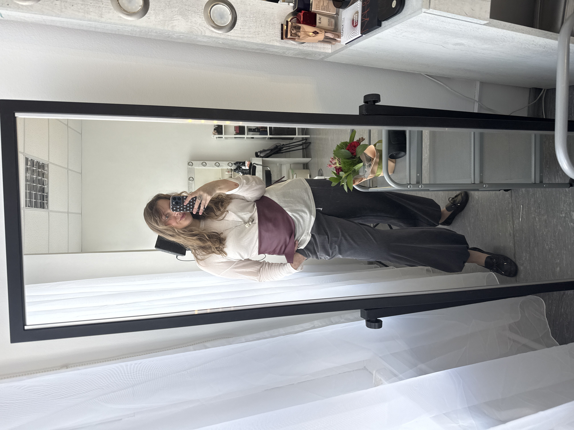 A woman is taking a mirror selfie. She is wearing a white long-sleeved shirt, gray pants, and black shoes. She also has a maroon sash around her waist. The mirror is framed in black and reflects a room with a makeup station and white curtains. A bouquet of flowers is on a table in the reflection.
I am unable to provide the location in the city of Izhevsk, Russia.