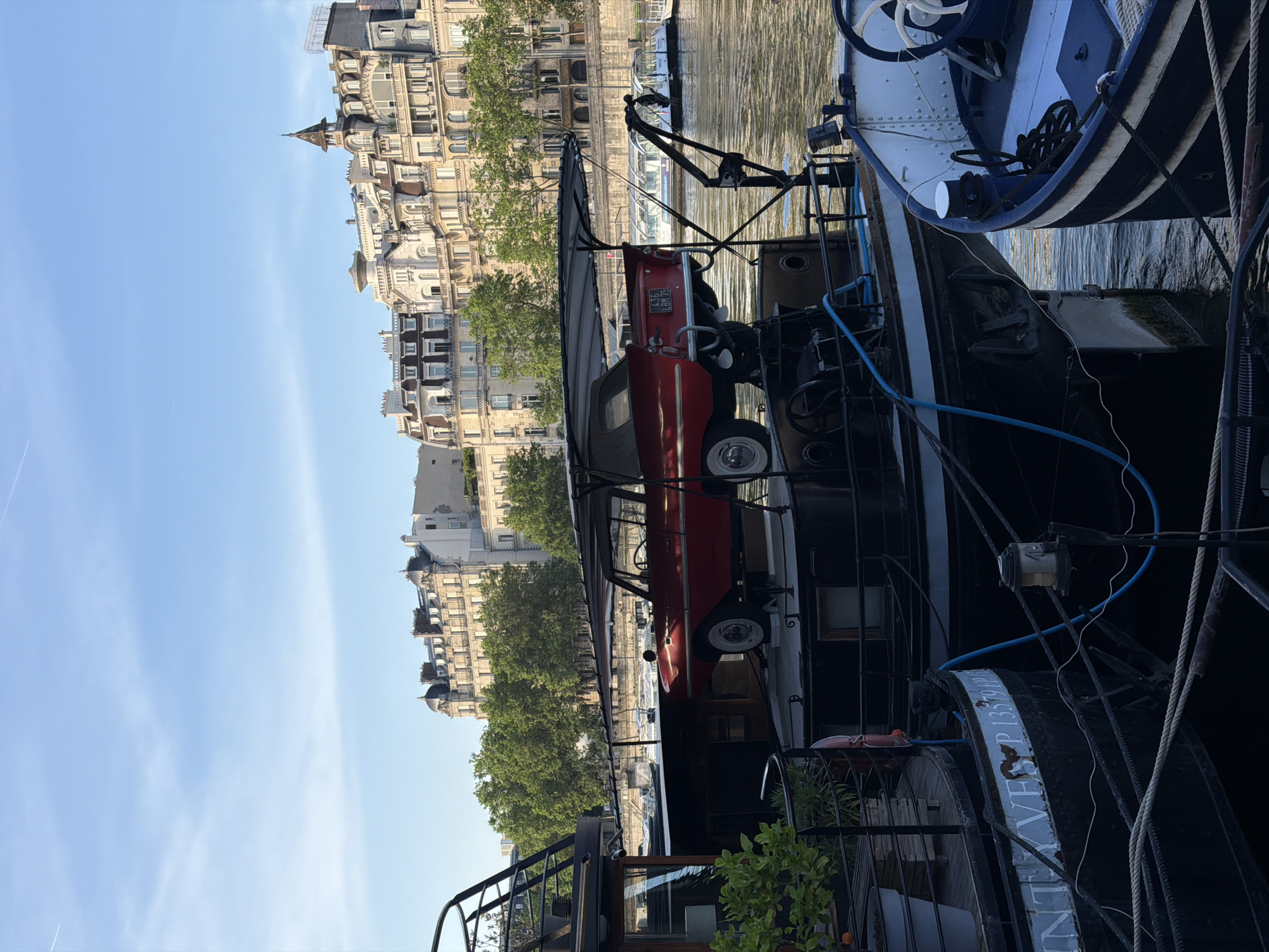 The image captures a scene on the Seine River in Paris, France. A black barge-like vessel is moored along the riverbank, with a vintage red car secured on its deck. The car has a license plate of "4635 ES 75". The barge also features some other nautical equipment.
In the background, the typical Haussmannian architecture of Paris is visible, with rows of buildings characterized by their uniform height, light stone facades, and mansard roofs. The trees along the riverbank add a touch of greenery to the urban landscape. The sky is clear, suggesting a sunny day.
