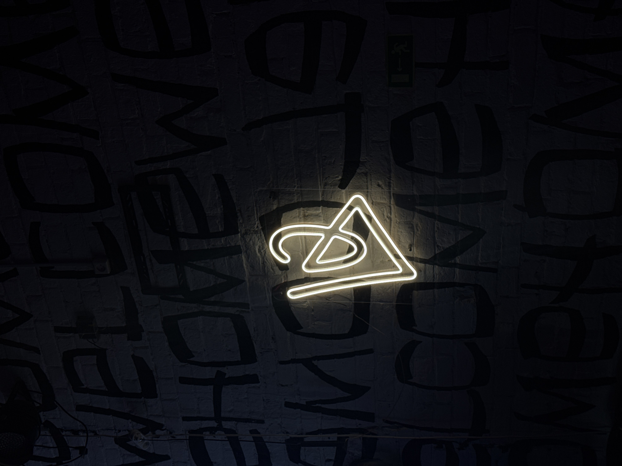The image captures an indoor space with a white neon sign resembling a stylized "ZA" or a combination of geometric shapes, possibly in the city of Perm, Russia. The sign is mounted on a dark wall with graffiti-style lettering, the word "WELCOME" being legible in several places. The wall appears to be made of brick. The lighting is dim, except for the bright neon sign, and an emergency exit sign is visible in the upper right quadrant.
