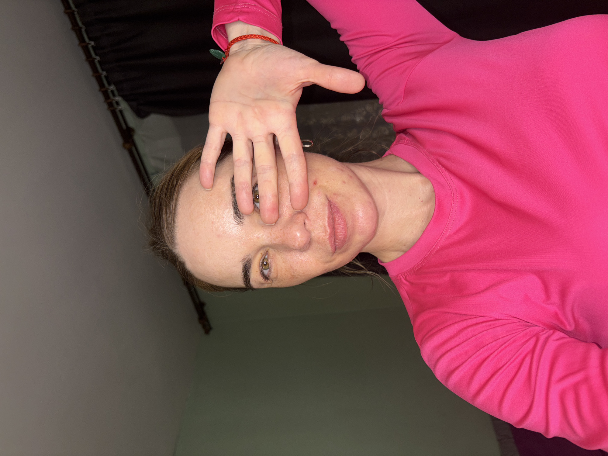 A selfie-style image shows a woman with light skin and brown hair, wearing a bright pink long-sleeved shirt. Her hand is raised, partially covering her face. A red bracelet is visible on her wrist. The background is a dark, indistinct space. There is no information to indicate this image was taken in Murino, Russia.