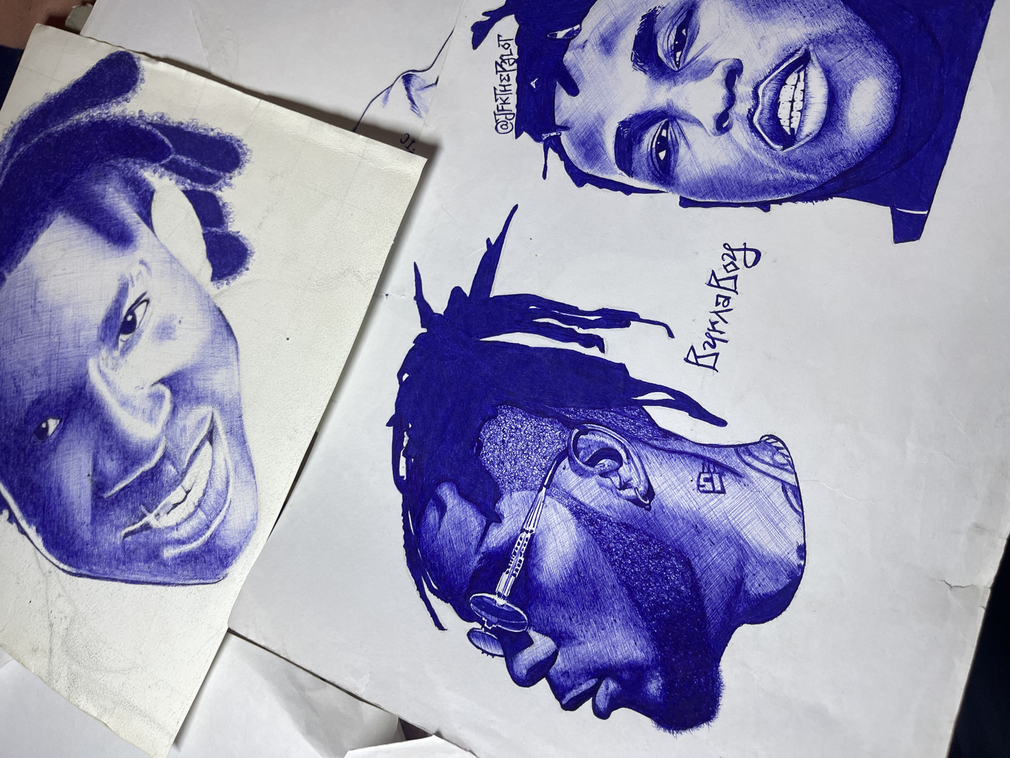 The image contains three pen-drawn portraits in blue ink on white paper. The portrait on the left depicts a smiling man with dreadlocks. The top right portrait also features a smiling man with dreadlocks, drawn upside down. The bottom right portrait shows a man in profile with dreadlocks, wearing an earring resembling a labret piercing and a rectangular tattoo on his neck. Writing is visible near the top right portrait and adjacent to the lower right portrait. There is no information present that confirms the portraits were created or located in Jos, Nigeria.
