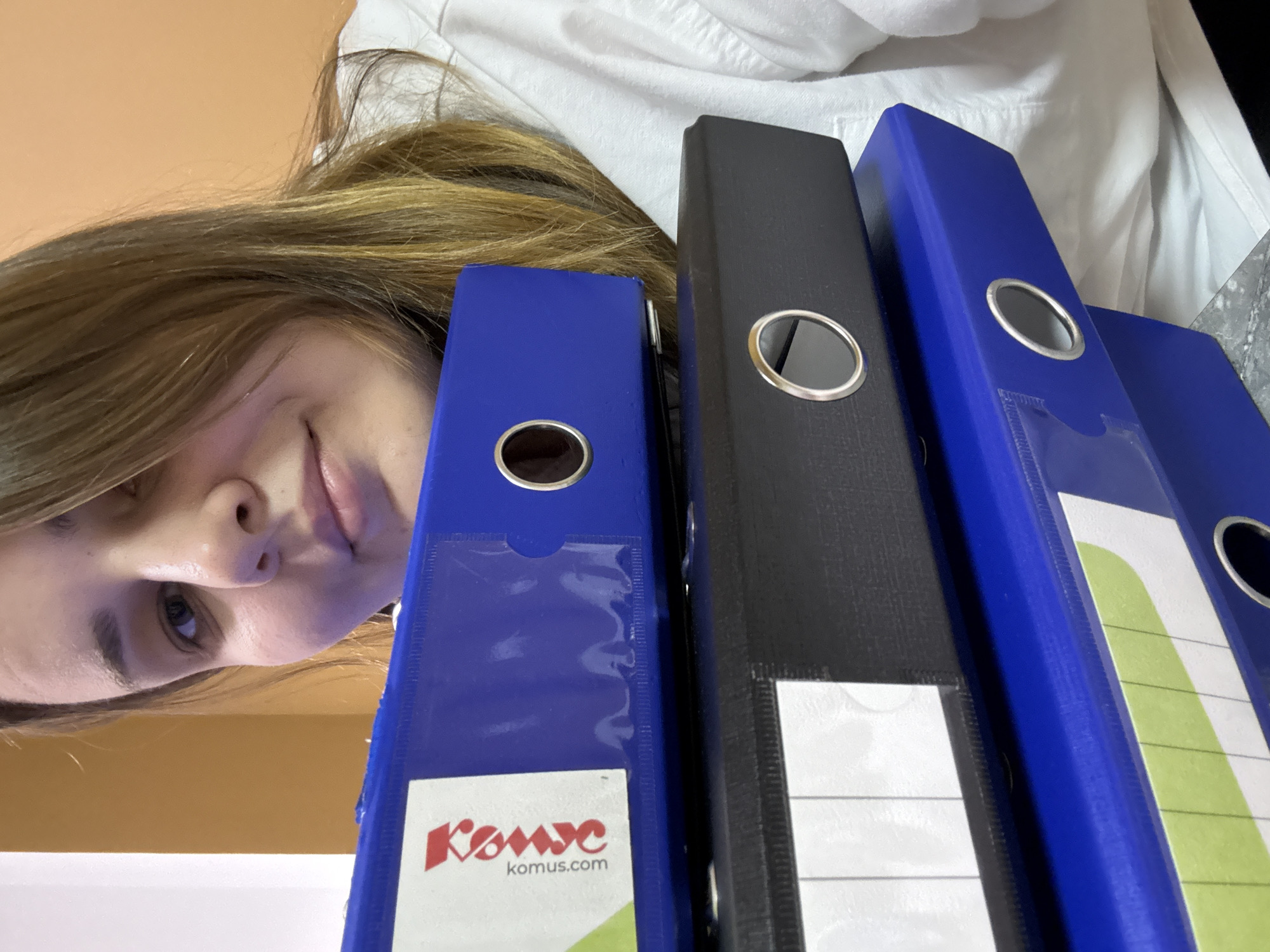 An eye-level shot captures a young woman looking down at a row of office binders, taken from an upward perspective in an indoor setting in Chaikovsky, Russia. Her face, framed by light brown hair, is partially visible in the upper left quadrant, exhibiting a subtle, pleasant smile. She appears to be wearing a white shirt, parts of which are visible behind the binders.
Three prominent ring binders are aligned vertically in the center and right of the frame: two blue and one black, with another partially visible blue binder to the far right. Each binder features a metal finger-hole ring and a clear plastic label holder. The leftmost blue binder clearly displays the red 'Komus' logo and 'komus.com' website on its label. The black binder and the rightmost visible blue binder have white labels, some with green and white striped patterns, and others blank. The woman seems to be observing or interacting with the binders, suggesting an activity related to organizing or working with documents. The background includes a plain light orange wall behind her head on the left and a textured white fabric, possibly part of her clothing or another item, behind the binders. There are no clear indicators of the time of day or weather.