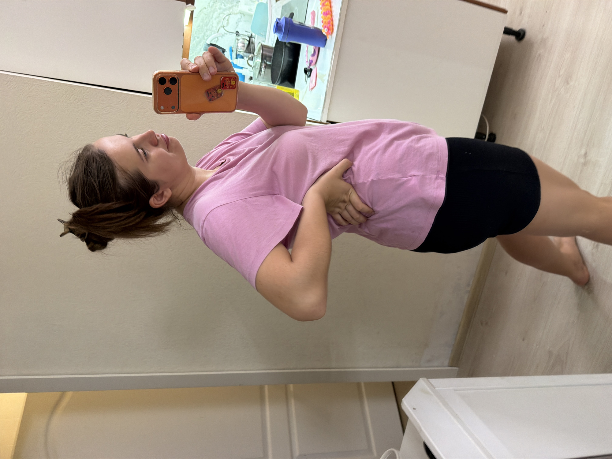 The image displays a woman taking a mirror selfie in an indoor setting, likely a home in Sochi, Russia. She is depicted standing, looking towards her smartphone, which she holds up to capture her reflection. The woman has light brown hair styled in a messy bun and is dressed in a light pink short-sleeved t-shirt paired with black shorts. Her left hand rests on her stomach area.
She is holding a smartphone encased in a vibrant orange cover, which features a prominent camera module and a few small, colorful cartoon-like stickers on its back. The reflection in the mirror behind her reveals what appears to be a kitchen or a utility space, with various items including a blue shaker bottle, pots or pans, and other kitchen essentials visible on shelves or a counter.
The room features light-colored walls and light wooden flooring. A white cabinet or piece of furniture is partially visible on the right side of the frame, with a white door or wall panel on the left. The lighting in the scene suggests an indoor environment, likely illuminated by artificial light. There is no discernible text visible within the image.