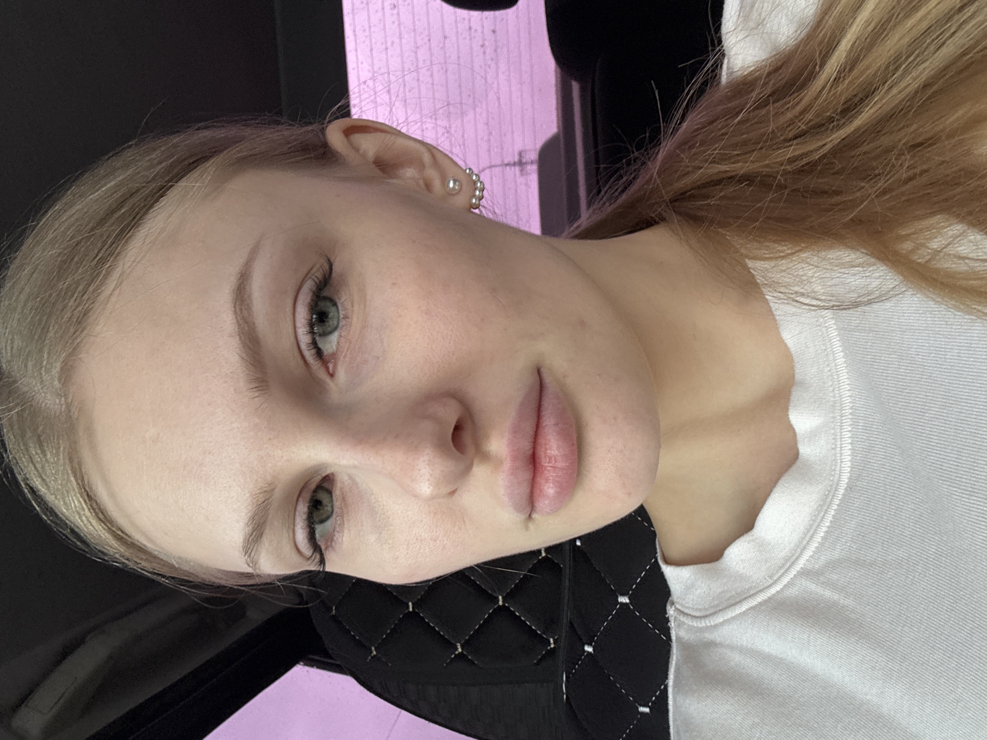 A close-up photograph depicts a young, fair-skinned person with light blonde hair partially pulled back. They have light-colored eyes, full lips, and are wearing an earring consisting of multiple small pearls in a row. The person is wearing a white t-shirt and is situated on what appears to be a black quilted material. The background includes a lavender or pinkish hue, possibly from interior lighting or reflections. The location cannot be determined to be Perm, Russia from the visual information in this image.