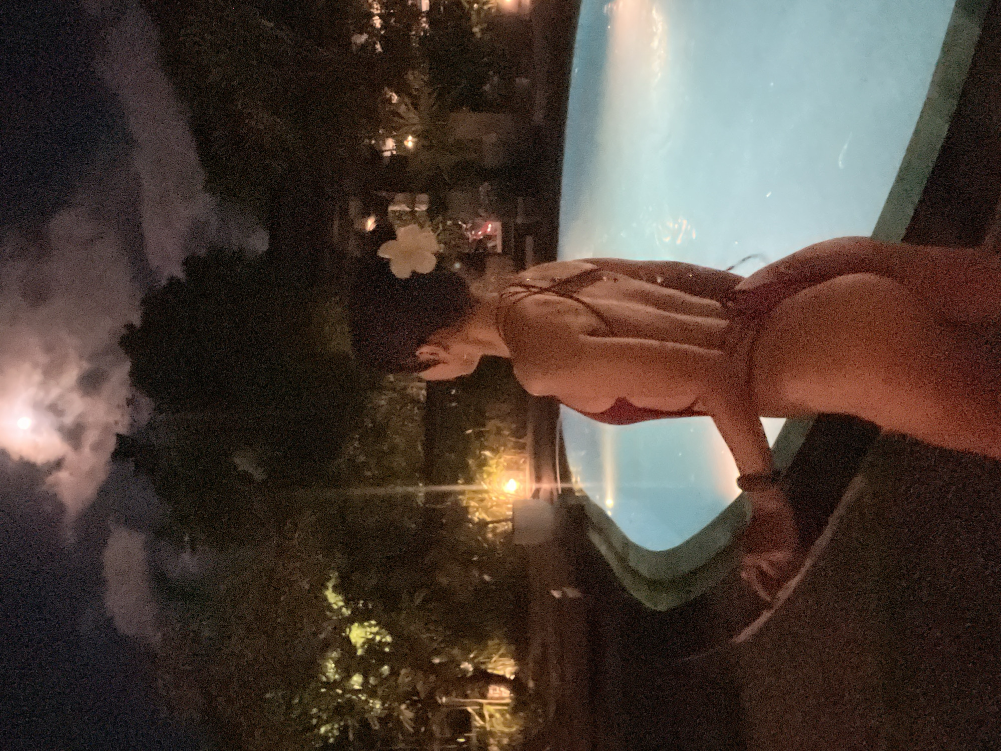 This nighttime image, captured in Kuta Selatan, Indonesia, shows a person leaning over the edge of a swimming pool. The subject, viewed from the back and side, is wearing a dark-colored swimsuit and has a white flower tucked into their hair. Their body is partially illuminated by ambient light.
The swimming pool, filled with light blue water, occupies the foreground and midground, reflecting nearby light sources. The water appears calm with subtle ripples. The background is dark, showcasing dense foliage and trees that line the pool area. Scattered among the trees are several warm-toned artificial lights, casting soft glows and creating depth in the scene. In the upper left corner of the dark sky, a bright, full moon is visible through wispy clouds, contributing to the overall illumination. The atmosphere is serene and suggests a tropical or resort setting. No text is visible in the image.