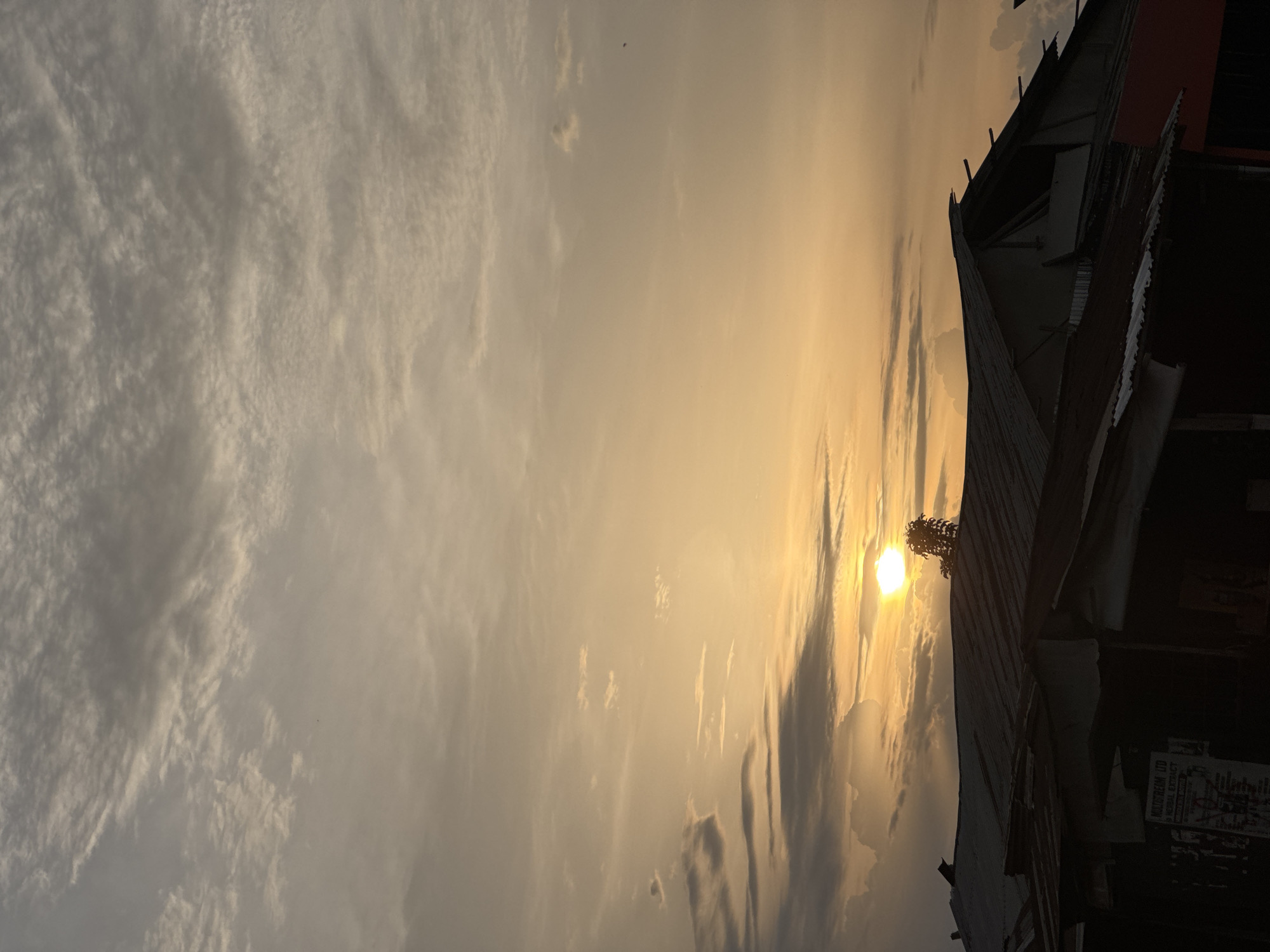 The image captures a vibrant sunrise or sunset over a local settlement in Ihiagwa, Nigeria, viewed from a low angle. The sky dominates the frame, showcasing a dramatic interplay of light and clouds. The sun, a brilliant golden orb, is positioned towards the right side of the frame, partially obscured by clouds and the dark silhouette of a foreground structure.
The sky is a tapestry of warm hues, with intense golden and orange light emanating from the sun and gradually fading into softer yellows and light greys towards the left. Various cloud formations are visible; some are wispy and scattered, while others are denser and appear darker against the bright light, casting subtle shadows across the sky.
In the foreground, along the bottom right edge, dark, silhouetted structures are visible, likely buildings or houses. These structures feature simple architecture, and what appears to be corrugated metal roofing can be discerned on some. A pole or antenna-like object rises from one of these roofs, close to the sun's position. Sparse, dark foliage, possibly from a small tree or bush, is also visible near the sun atop a roof.
The time of day is clearly either sunrise or sunset, characterized by the low angle of the sun and the warm, golden glow that bathes the entire scene, creating strong contrasts and silhouetting the foreground elements. The weather appears partly cloudy. No people are visible.
A notable detail is a faded sign or poster on a dark wall of one of the buildings in the lower right corner. Text is partially legible, including "MULTISTREAM LTD" and "MINERAL EXTRACT", providing a subtle glimpse into local commerce or industry. The overall mood conveyed by the image is one of peaceful natural beauty and the serene close of day or dawn.