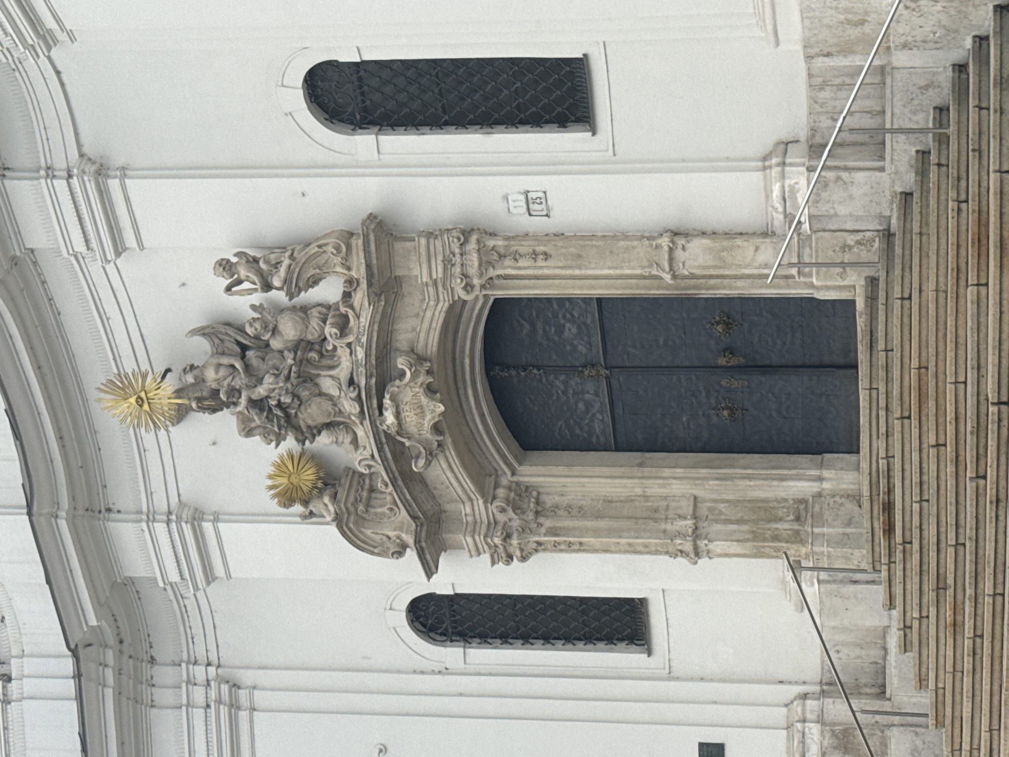 This image depicts a detailed view of an ornate architectural facade, likely a church or significant public building, in Bratislava, Slovakia. The primary subject is the elaborate stonework surrounding a large, dark double door. Flanking the doorway are decorative carvings of angelic or cherubic figures, along with other mythological or allegorical sculptures. Two starburst-shaped, golden ornaments are prominent near the top left of the sculpted section, adding a touch of opulence.
Above the main doorway, a smaller rectangular window with a decorative iron grille is visible, mirroring a similar window on the lower left of the image. A plaque with the numbers "11" and "L25" is mounted on the wall above the doorway. The stonework itself is a mixture of light and weathered grey stone, showing intricate carvings and moldings. To the right of the doorway, a section of a grand, tall staircase constructed from vertical stone slabs is visible, suggesting a monumental entrance. The overall aesthetic is one of historical grandeur and religious or civic importance. The lighting suggests it is daytime, with no clear indication of weather conditions.