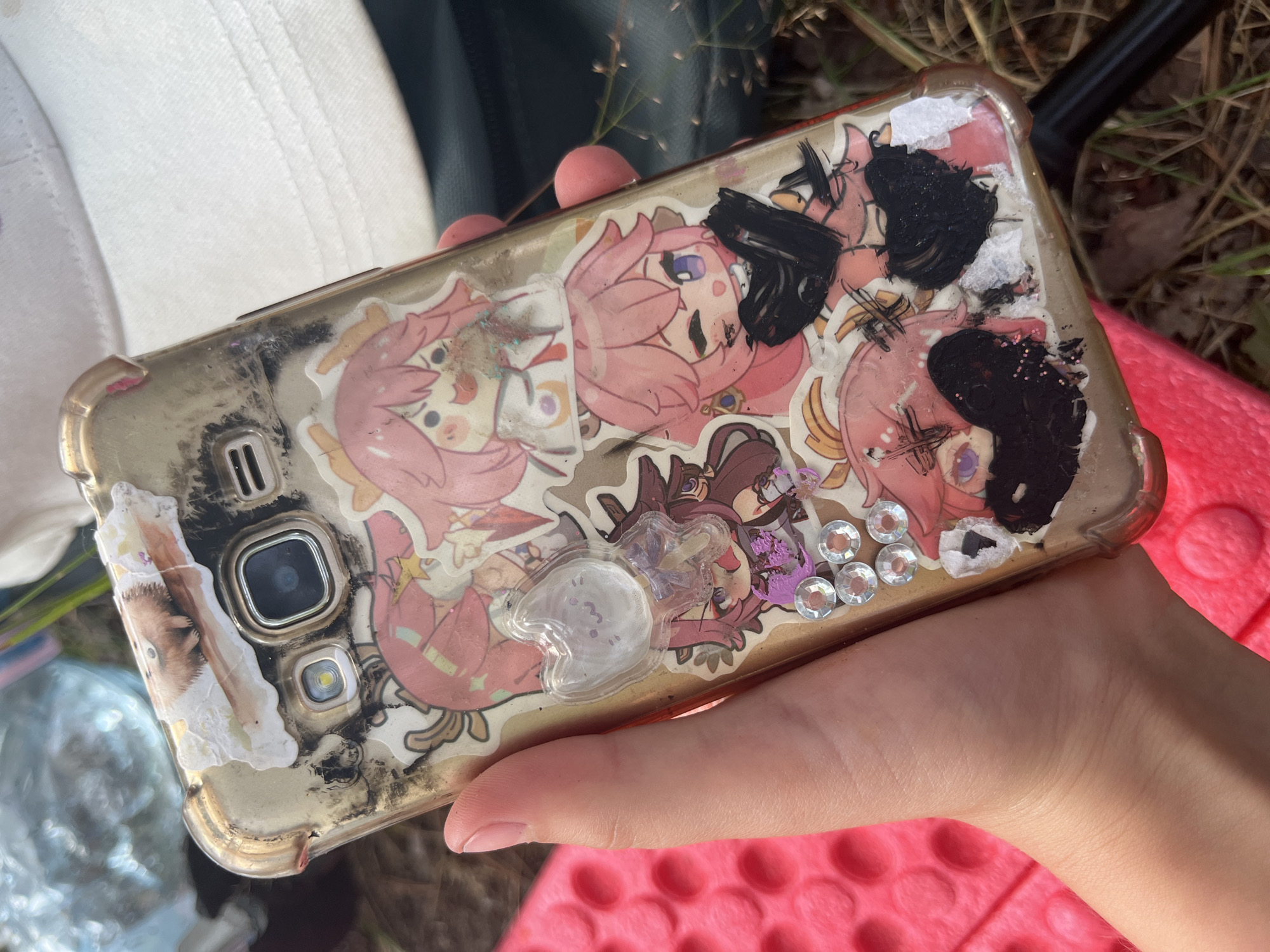 The image shows a person's hand holding a gold-colored phone case decorated with anime-style stickers. The stickers are of characters with pink hair and large eyes. Some of the stickers appear to be damaged, with peeling edges and smudges of black material. There are also several small, clear rhinestones glued to the case. The phone case itself appears worn and scratched. In the background, out of focus, are hints of grass, a pink textured object, and a white hat. The image does not provide any information about the location in the city of Pillapalu, Estonia.