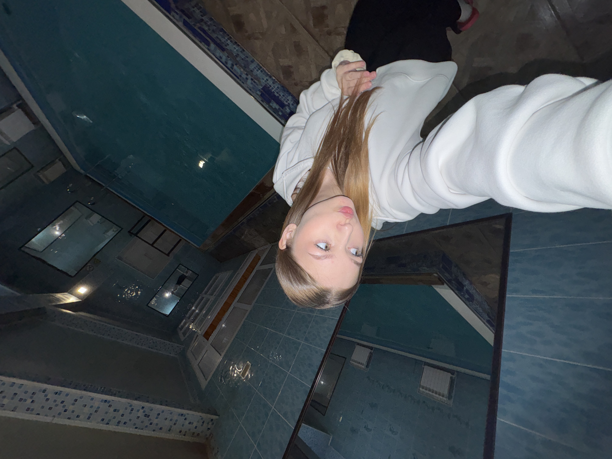 A woman with fair skin and blonde hair is taking a selfie in a tiled room with a pool. She is wearing a white hooded sweatshirt. The room is primarily tiled in a bluish-green color, and there is a pool with a white edge. Several windows are visible in the background. The location is Chaikovsky, Russia.