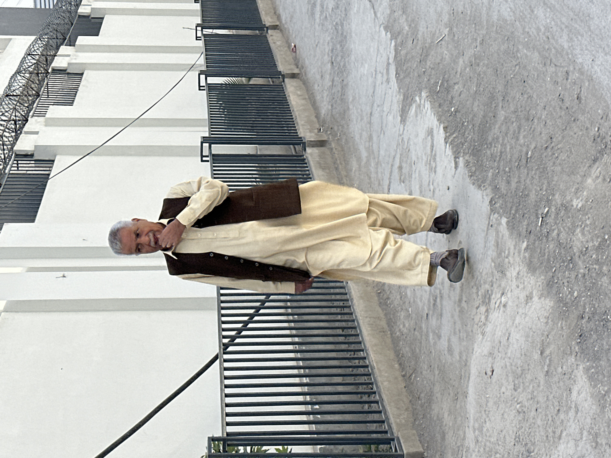 A man stands on a grey concrete surface next to a white building with security fencing, likely in Quetta City Tehsil, Pakistan. He wears a light yellow shalwar kameez with a dark brown waistcoat, and brown shoes. He is looking at the camera and has his hand raised to his mouth.