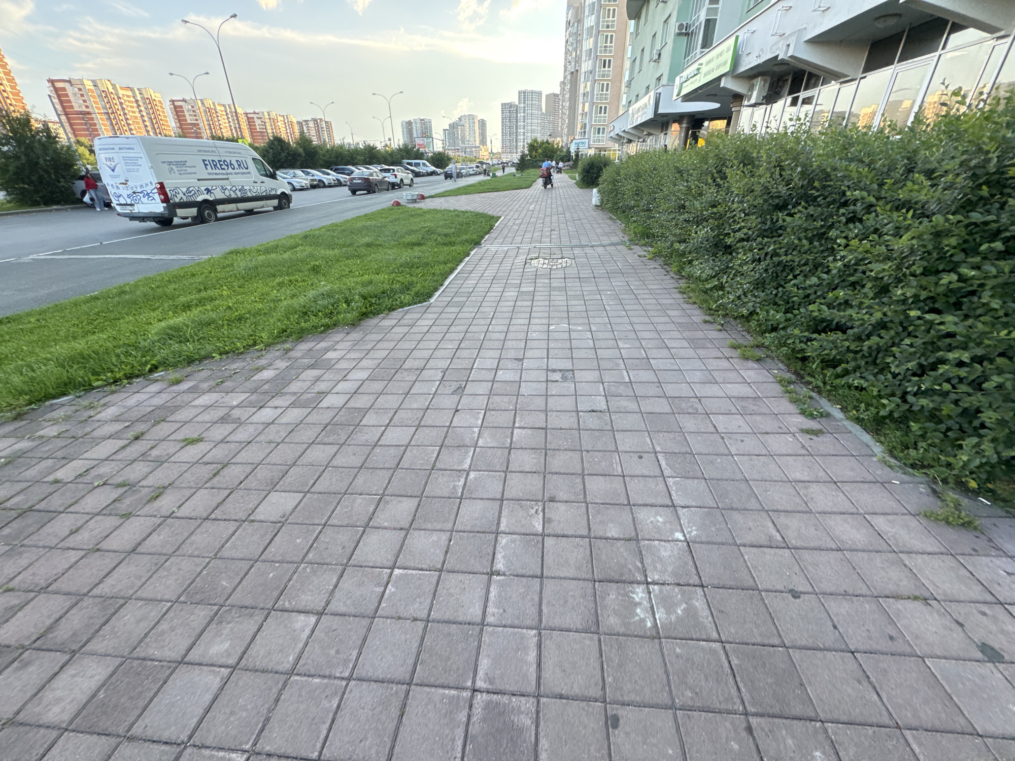 The image captures a street scene in Yekaterinburg, Russia. The foreground is dominated by a gray tiled sidewalk, with patches of grass and weeds growing through the cracks. To the left of the sidewalk is a strip of vibrant green grass that borders a road. Beyond the road, a white delivery van with "FIRE96.RU" prominently displayed on its side is parked. The van also features various graphics and text in Russian. Further down the road, a line of parked cars is visible, and in the distance, a row of modern, multi-story apartment buildings lines the street.
On the sidewalk, a person is riding a scooter in the middle distance, heading away from the camera. The setting appears to be an urban environment during the day, with a moderately cloudy sky. The architecture of the buildings suggests a contemporary cityscape. The overall impression is that of a typical street scene in a Russian city.