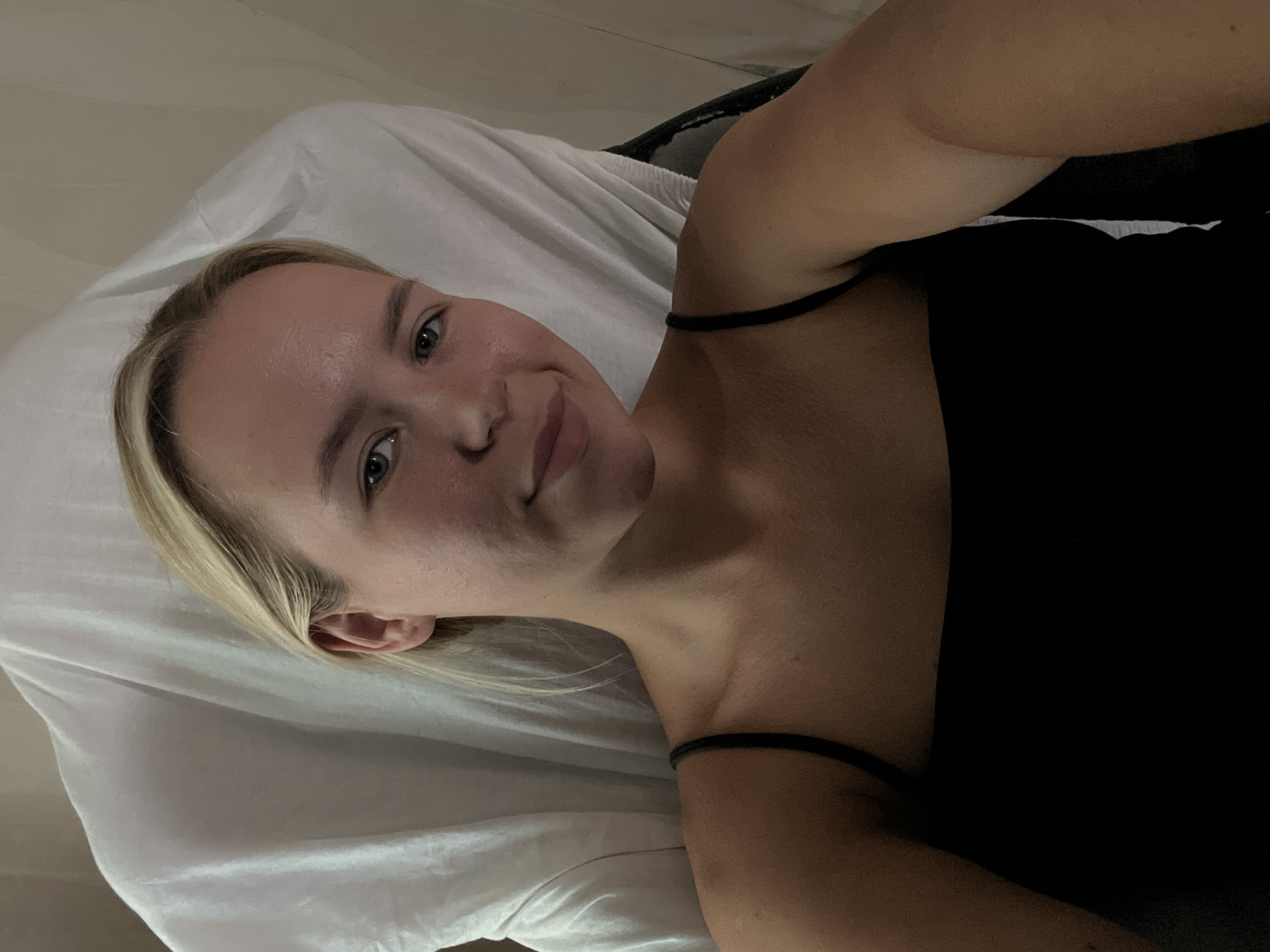 A young Caucasian woman with blonde hair and blue eyes is pictured from the chest up, looking directly at the camera. She is wearing a black tank top. Her head is tilted slightly to her left, and she has a slight smile. Behind her, a white sheet is draped over what appears to be a chair or bed. The lighting is dim, casting shadows on the right side of her face and body. The background is blurred and appears to be indoors, possibly in a bedroom or spa setting, in an unknown location.