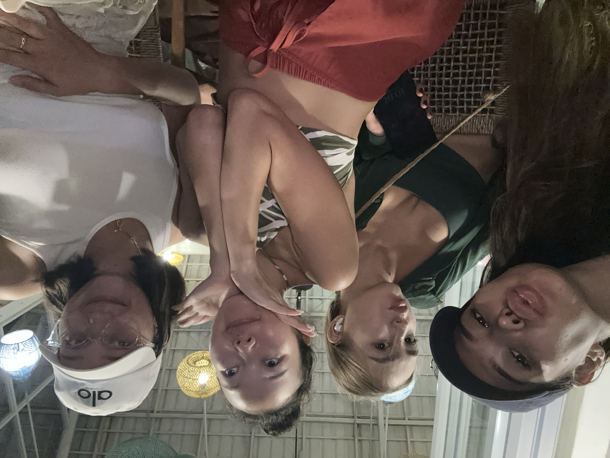 Four women are posing for a selfie in what appears to be an indoor or semi-outdoor venue, possibly in North Kuta, Indonesia. The woman on the left wears a white top, glasses, and a white baseball cap with "alo" written on it. Next to her, a woman is dressed in a bikini top with a leafy green and white pattern, paired with loose red shorts. A third woman to the right wears a green top. On the far right, the last woman wears a dark-colored baseball cap. Behind them, there is a lattice-like structure that may be part of the building's architecture. Lighting fixtures hang from the ceiling. They appear to be seated in an area with a woven surface.