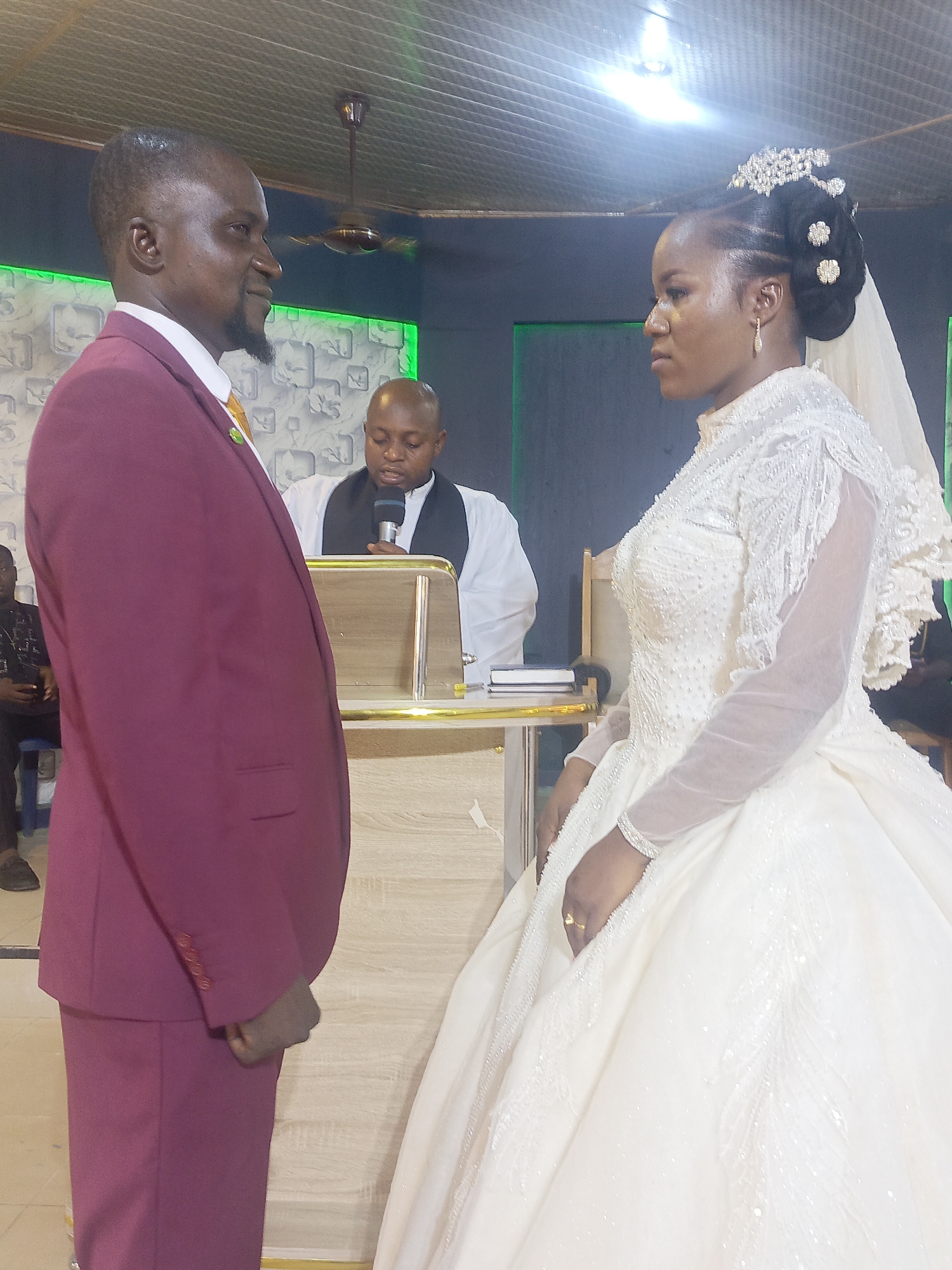 A wedding ceremony takes place in what appears to be a church or hall in Unknown, Unknown. The groom, dressed in a burgundy suit with an orange tie, stands to the left, facing the bride. The bride is adorned in a full-length, white wedding gown with embellishments, and a veil is attached to her hair, which is styled with an ornate hairpiece. An officiant stands behind a wooden podium between the couple, holding a microphone and presumably conducting the ceremony. The background includes decorative wallpaper, green LED lighting, and a ceiling fan. A few seated individuals are partially visible in the background. The setting suggests a simple, indoor wedding event.