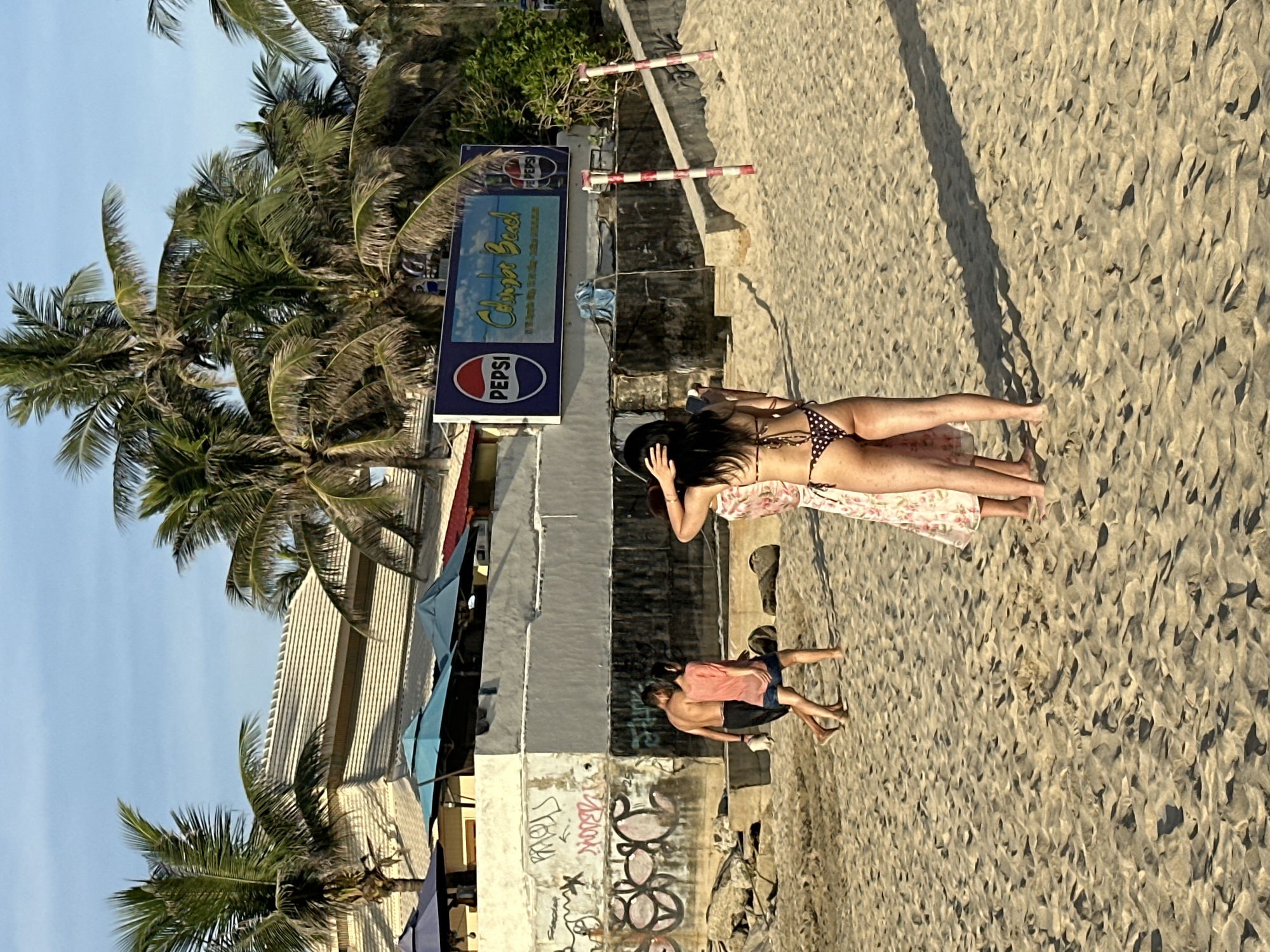 The image depicts a beach scene in Đà Nẵng, Vietnam, with a building and palm trees in the background. A large Pepsi advertisement is visible on the building. In the foreground, several people are present on the sand. Two individuals are walking away from the camera, a shirtless man in dark shorts and a person in a coral-colored shirt and denim shorts. To the right, a person in a bikini with a polka-dot pattern is facing away from the camera, with another individual in a floral dress partially visible beside them. The beach is sandy, with footprints and shadows indicating the time of day is likely late afternoon or early evening. The environment appears to be a tourist area, with umbrellas visible near the building.