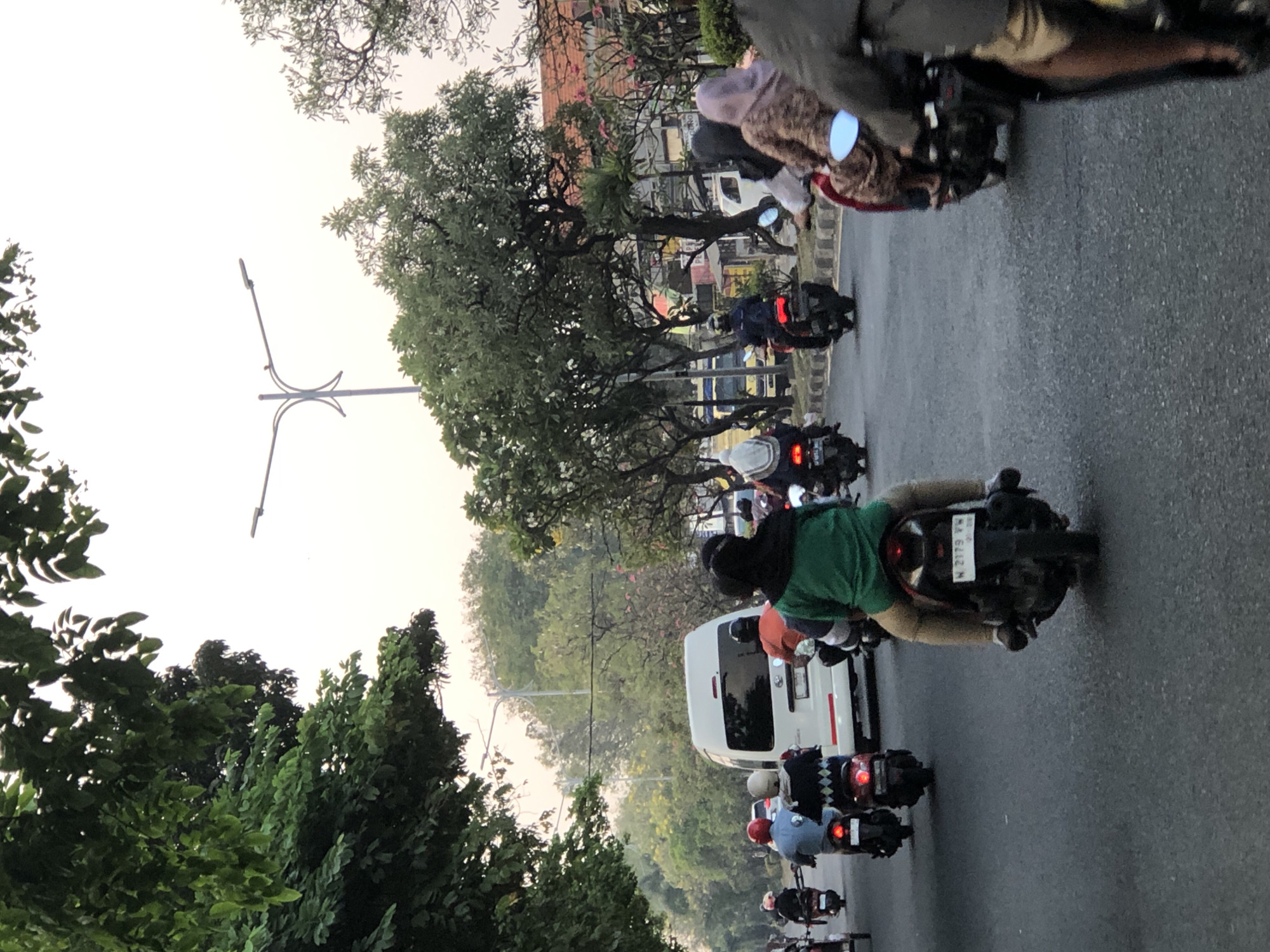 A traffic scene on a paved road with multiple motorcycles and a white van. The sky is overcast. Trees line the left side of the road, with some vegetation and buildings visible in the background on the right. Several individuals are riding on motorcycles, some with passengers. The motorcycle in the foreground on the right has a rider wearing a dark jacket and tan pants, and the license plate is partially visible. Another motorcycle in the middle of the frame has a rider in a green shirt and tan pants, with a passenger behind. The white van is in the center, with other motorcycles and riders in front and behind it. The environment suggests a bustling urban or suburban setting.
