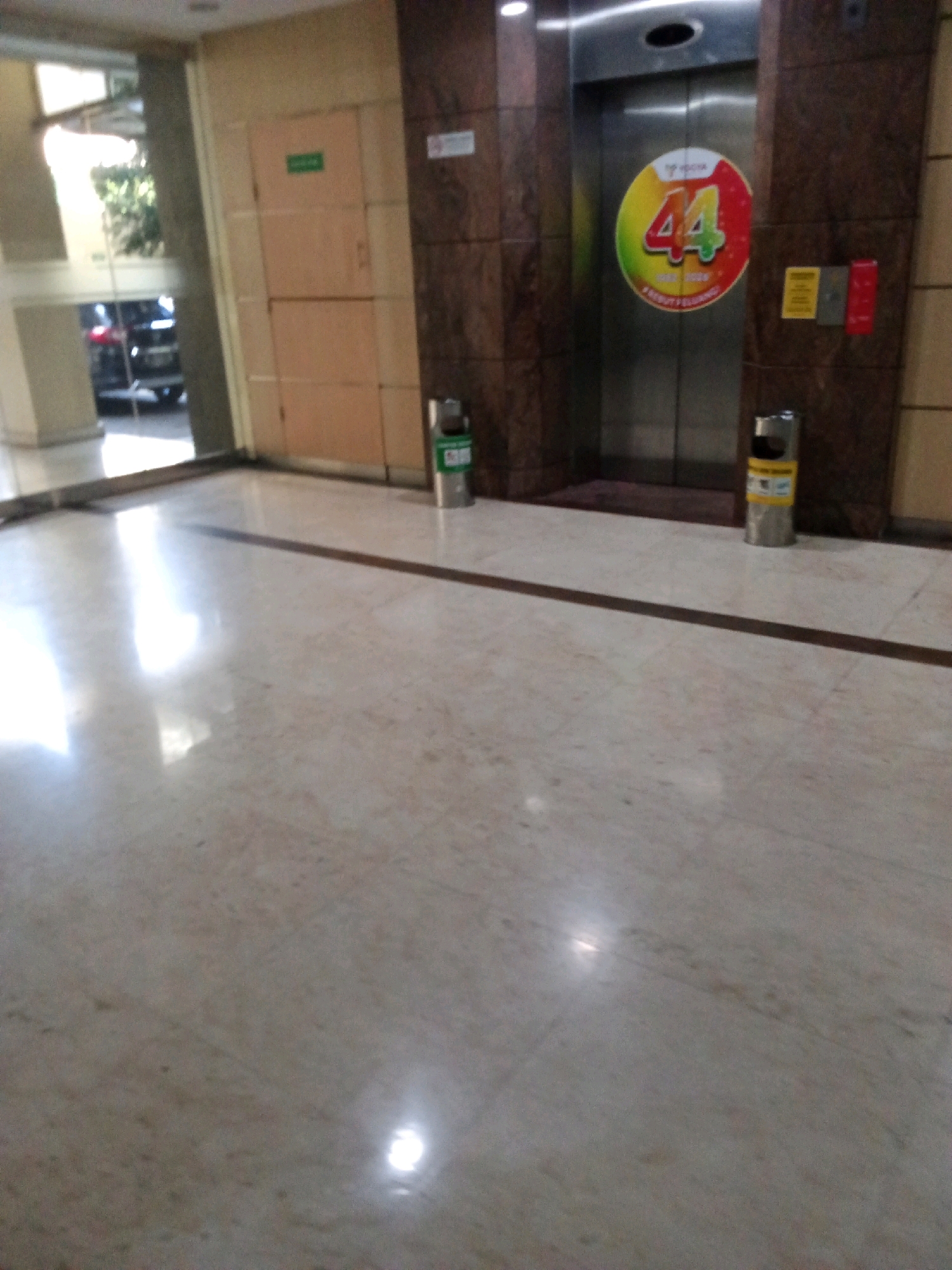 The image shows a hallway with a light-colored tiled floor and a dark brown stripe running horizontally across it. On the right side of the hallway, there is an elevator with a large circular sticker on its doors featuring the number "44" in red and green. Next to the elevator, there are two trash cans, one silver and cylindrical with a yellow label, and another smaller green one. To the left of the elevator is a door with a green sign. Further to the left, there is a doorway leading to an area with sunlight and a parked car. The environment appears to be an indoor public space, possibly a building lobby or corridor.