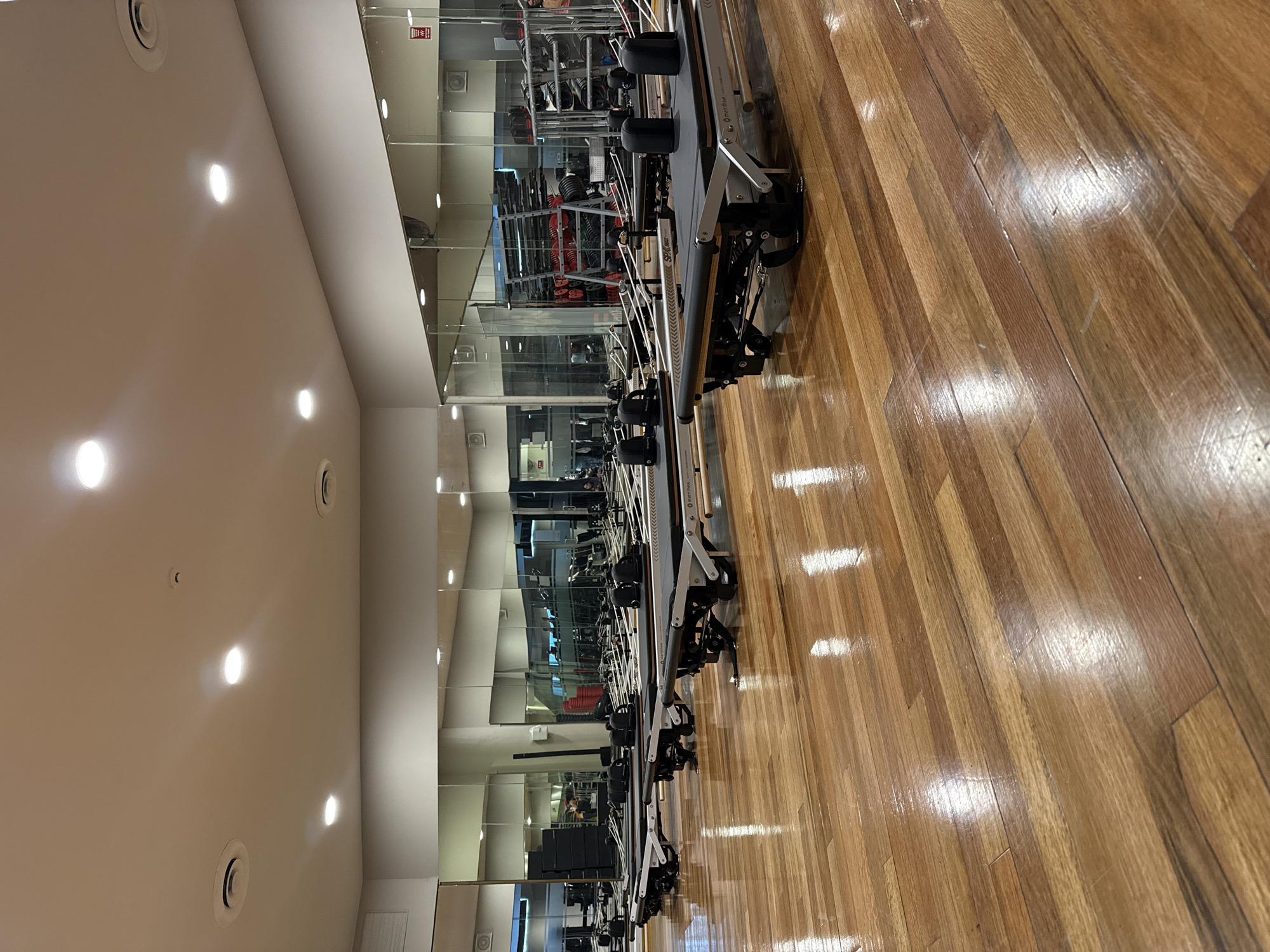 The image depicts an indoor exercise space with a row of Pilates reformer machines. The room has a wooden floor, mirrored wall, and a white ceiling with recessed lighting. The machines are aligned along the wall, with reflections in the mirrors showing additional gym equipment in the background. There is no visible human activity. There is nothing in the image to indicate that the location is in Puebla, Mexico.