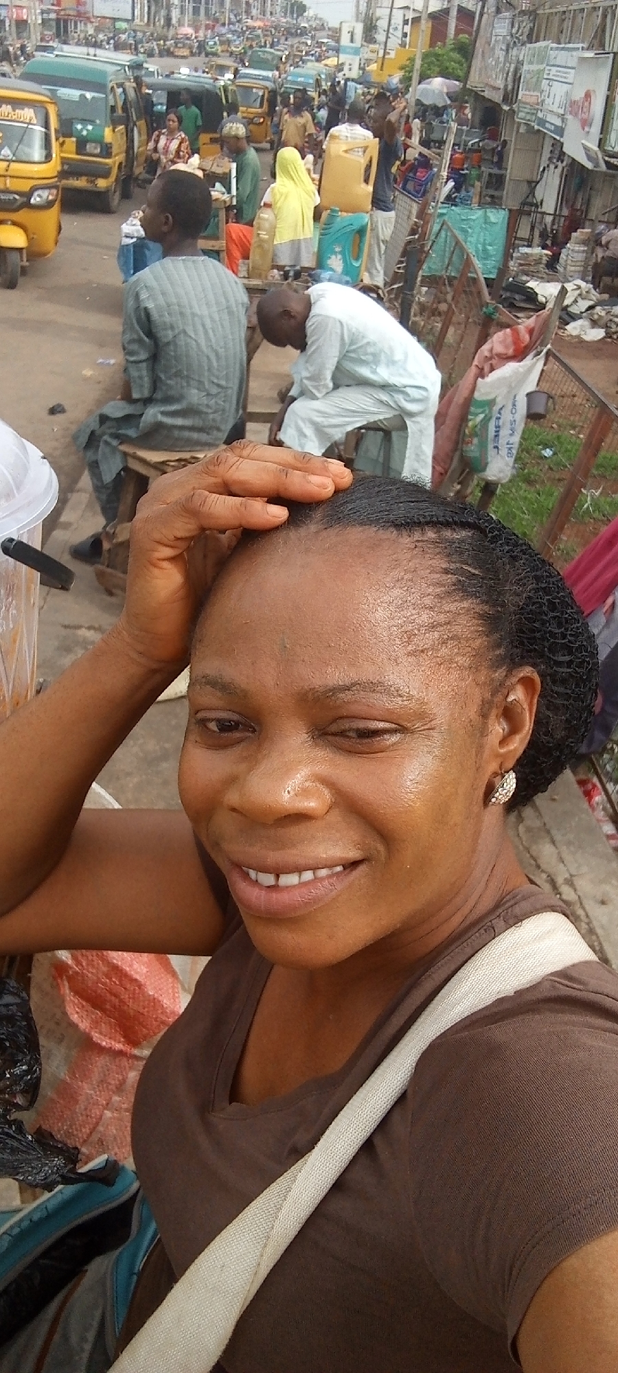 A woman with dark skin and a brown t-shirt smiles and touches her forehead. She stands on a sidewalk in Maiburiji, Nigeria. Behind her, a busy street scene unfolds, featuring yellow auto rickshaws, pedestrians, and people sitting on benches. The background shows buildings and various objects typical of a street market.