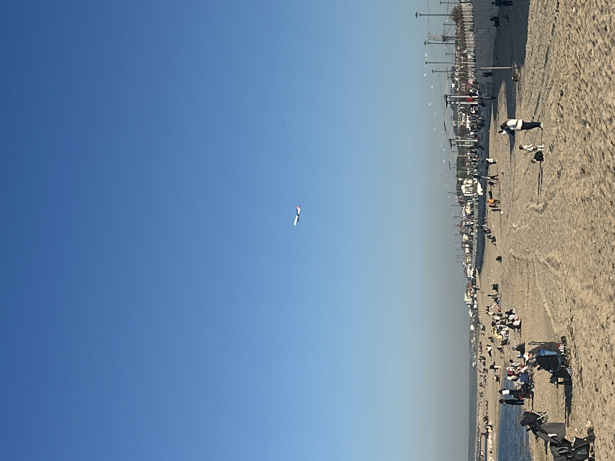 The image shows a beach scene on a sunny day. People are scattered across the sand, with some sitting, standing, or walking. The beach borders a developed area featuring buildings and lampposts. In the clear blue sky, a kite is flying. The location is Arnavutköy, Turkey.