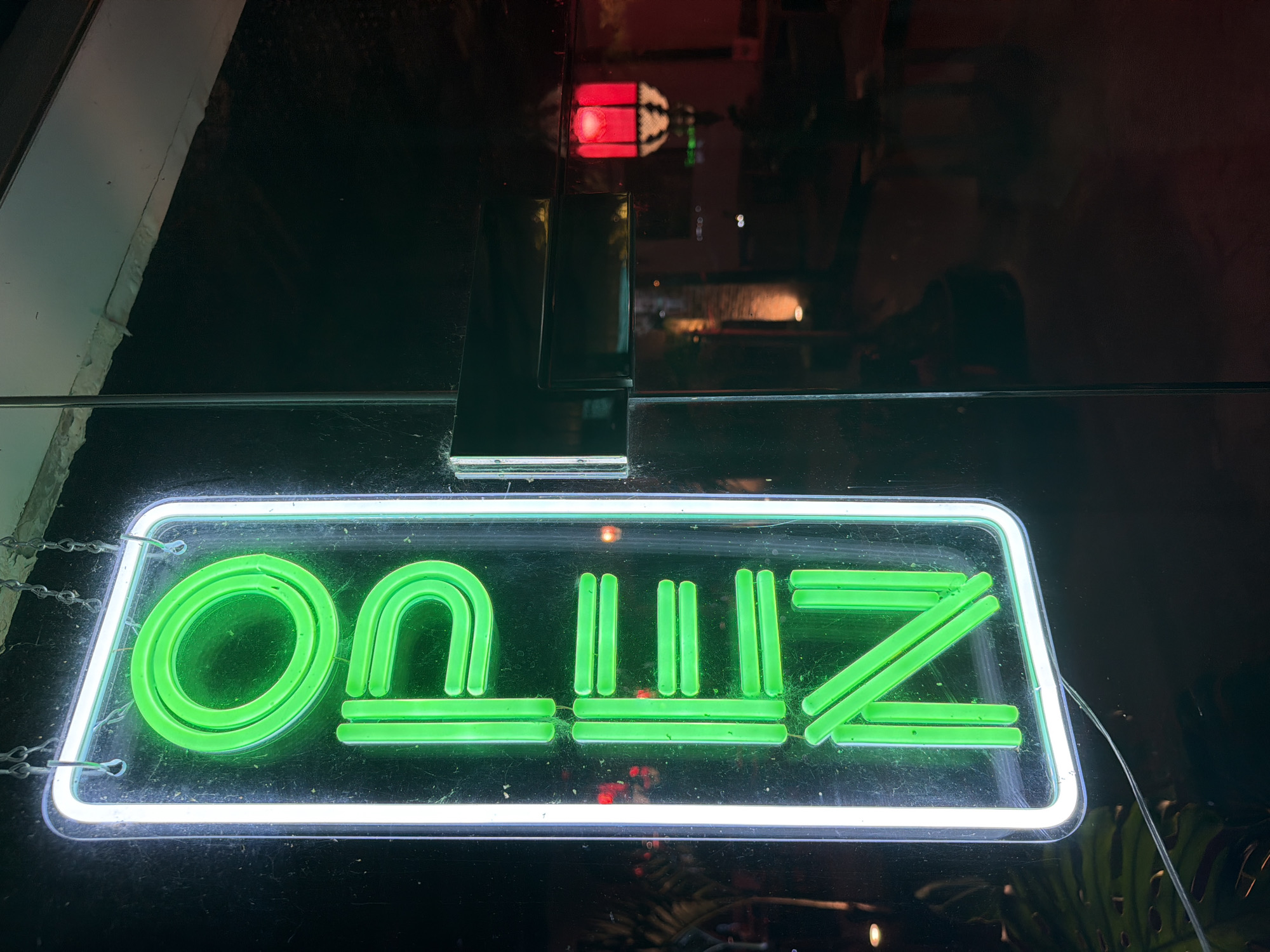 An illuminated "OPEN" sign, composed of green neon tubes within a white neon border, hangs vertically by chains from a ceiling. The sign is affixed to a glass surface, likely a storefront window, which reflects a dimly lit interior. The interior scene includes hints of red lighting and possibly furniture. The overall environment appears to be nighttime. The location is identified as Baan Bang Rak, Thailand.