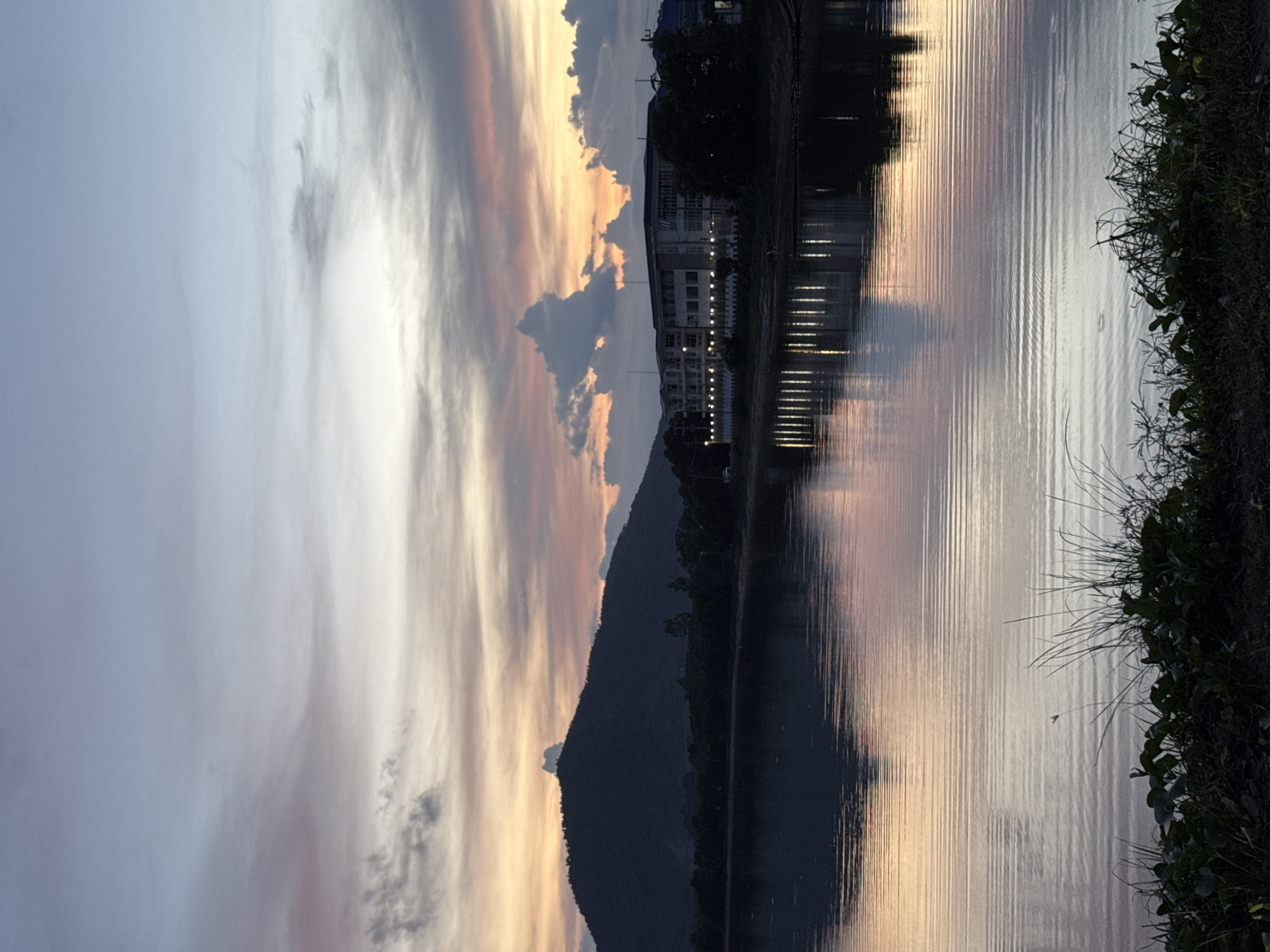 This is a landscape photograph taken at dusk or dawn, featuring a body of water in the foreground and a hill and buildings in the background. The sky displays a gradient of colors, with shades of blue, pink, and orange hues blending together, indicating either sunrise or sunset. The water reflects the sky and the structures on the opposite bank, creating a mirror-like effect with gentle ripples distorting the reflections. On the left side of the background, a dark silhouette of a hill dominates the horizon, its shape rounded and covered in vegetation. To the right of the hill, a multi-story building with numerous windows and balconies is visible, illuminated by lights, suggesting it is a hotel or residential complex. Trees and other foliage line the bank between the water and the building. The foreground shows the edge of the water with dark, silhouetted vegetation, including grass and broad leaves, growing along the bank. The overall mood of the image is serene and tranquil.
