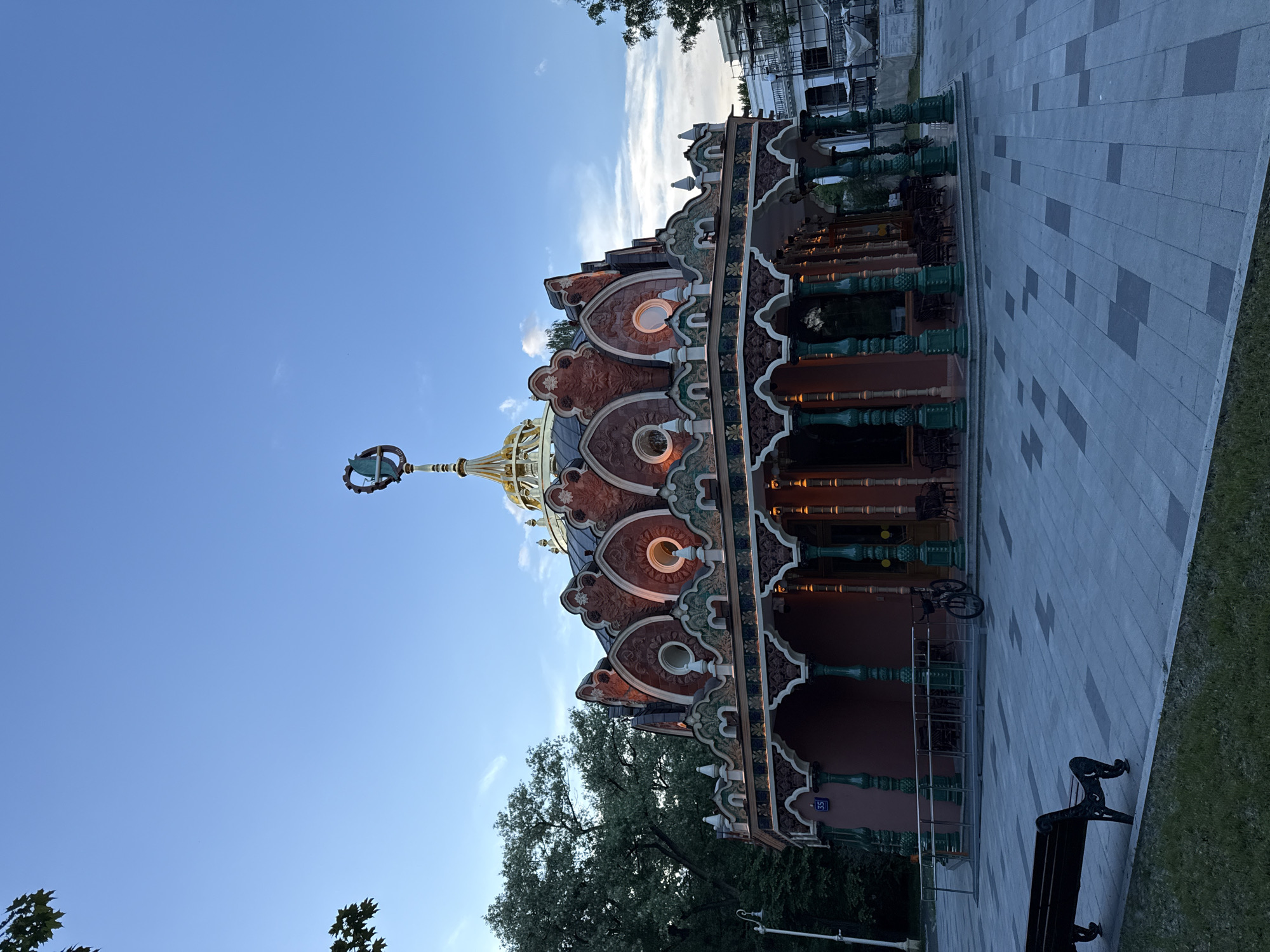 An ornate, multi-storied building with a distinctive golden dome and spire dominates the right side of the image, set against a bright, clear blue sky with sparse white clouds. The building features rich architectural details, including red and dark green facades, intricate patterns, and circular windows that appear to emit a warm glow. The overall design suggests a traditional Russian or Eastern European style, possibly a pavilion or a small palace within a park.
In the foreground, a wide paved walkway, made of light and dark grey rectangular tiles arranged in a patterned grid, leads towards the building. A classic black cast-iron park bench rests on the paved path, next to a small strip of green grass. A bicycle is parked near the building's entrance. Green trees are visible at the bottom left and partially behind the structure, hinting at a natural, park-like environment. In the background, beyond the main building, scaffolding or another building under construction can be seen.
The lighting indicates late afternoon or early evening, characterized by the bright sky and the warm, almost illuminated appearance of the building's details. No people are visible, contributing to a serene atmosphere. A small blue sign with the number "35" is visible on the side of the building. The scene depicts a tranquil urban landscape, likely within a park in Moscow, Russia.