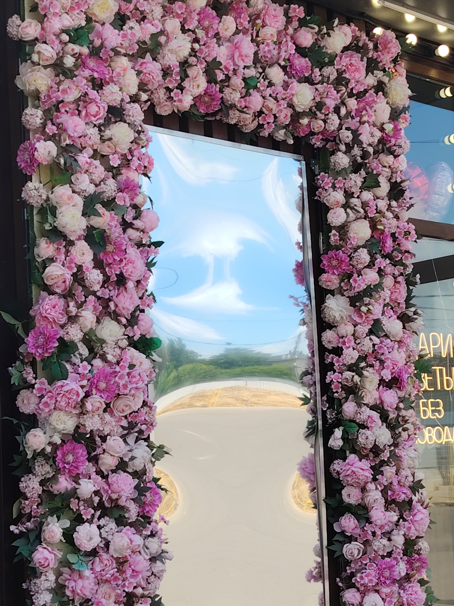 A floral archway constructed of pink and white artificial flowers frames a reflective surface, likely a mirror or a window, in front of a building. The reflective surface shows a distorted view of a blue sky with white clouds and a hint of greenery and a road. To the right of the archway, partially visible text in neon yellow lights is displayed on the building's facade. The text appears to be in Russian, with some legible words including "АРРИ" and "БЕЗ". The setting suggests an outdoor urban environment, possibly a shop entrance or a decorative installation in the city of Dolgoprudny, Russia, given the contextual information. No people are visible in the frame.