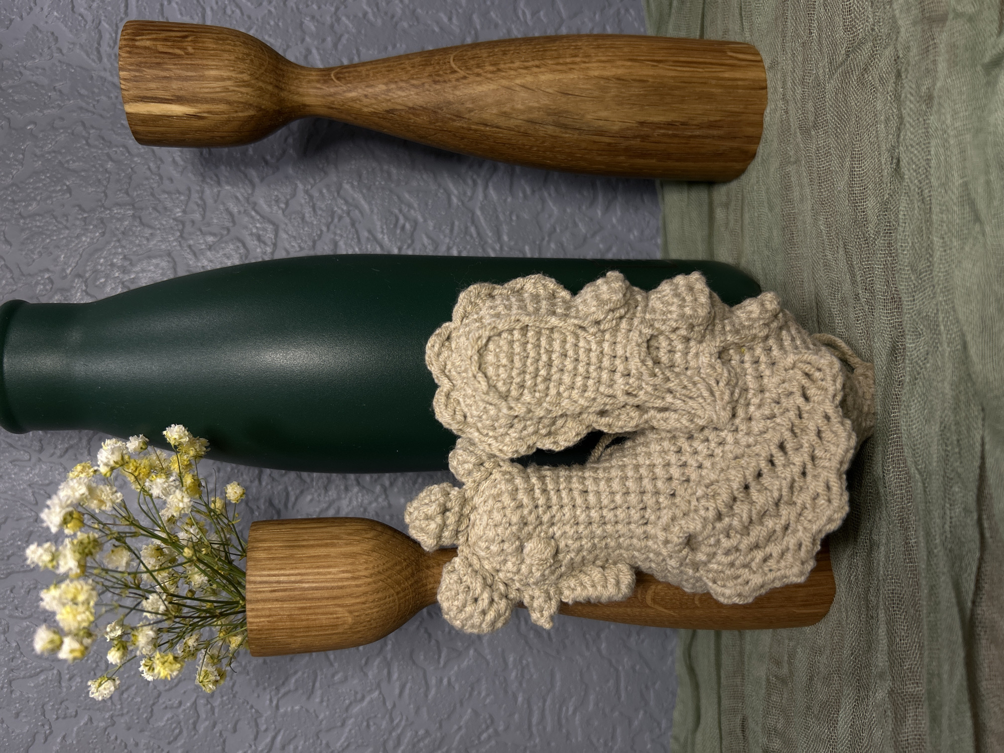 The image displays a flat lay still life arrangement, originating from Saint Petersburg, Russia. A dark green bottle or thermos, smooth and cylindrical, lies horizontally across the center of the frame. Above it, a wooden, spindle-shaped object tapers from a wider end on the left to a narrower end on the right, showcasing its natural wood grain. Below the bottle, another similar wooden object, wider on its right end, serves as a small vase holding a cluster of delicate white and pale yellow flowers, resembling baby's breath.
Draped over the green bottle and extending towards the lower wooden piece is a beige, crocheted item, possibly a decorative scrunchie or a small embellishment, featuring a distinct, slightly lumpy texture with ruffled edges. The objects are set against a textured grey background, reminiscent of concrete or stucco. To the right, a sheer, light green, loosely woven fabric adds another layer of texture and color to the composition. No people are visible, no specific activity is occurring, and there is no discernible text. The lighting suggests an indoor setting.
