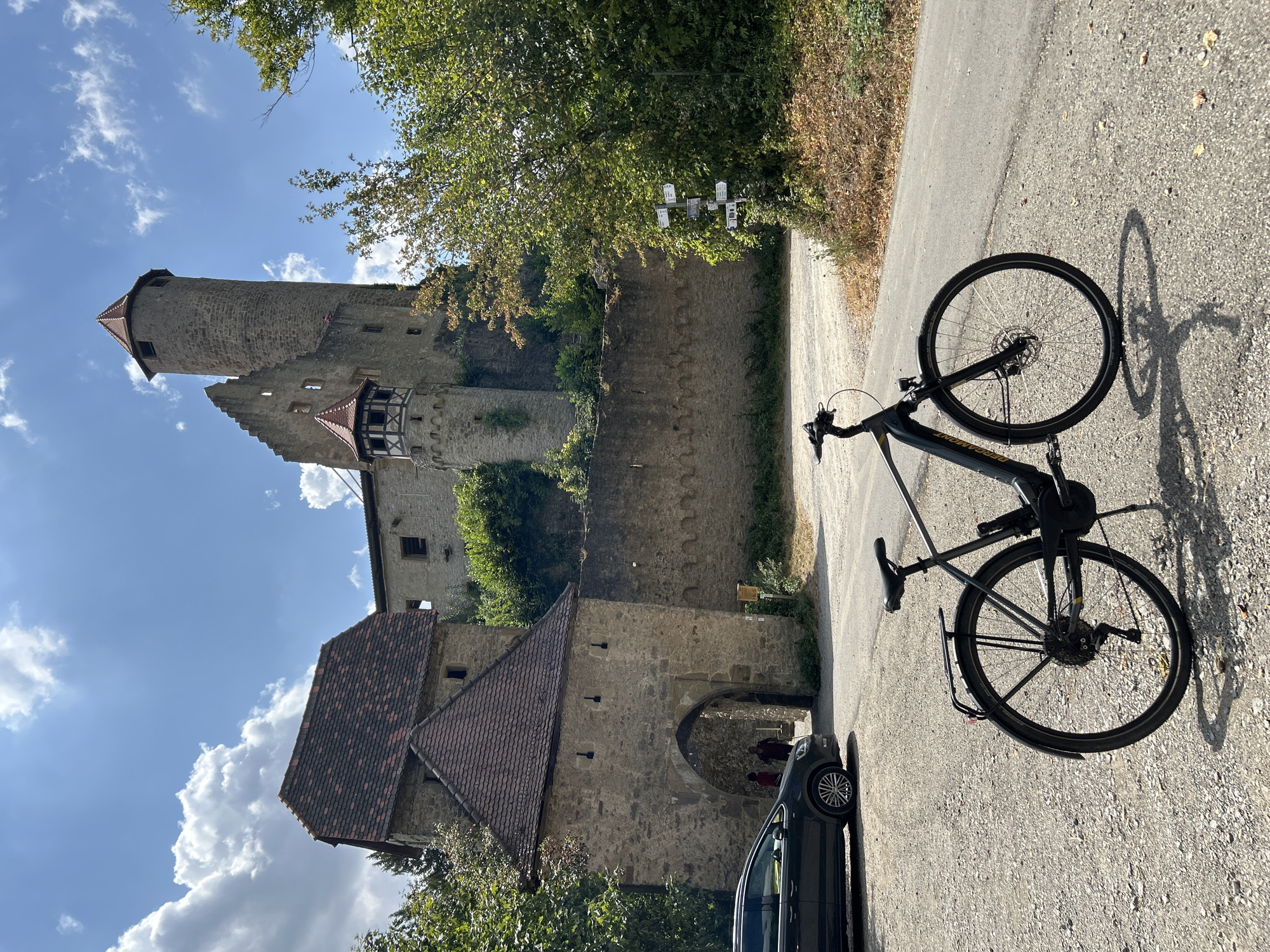 The image depicts an electric bicycle parked on a gravel path in front of an ancient stone castle complex. The castle, identifiable as Burg Hornberg in Neckarzimmern, Germany, features a tall, round tower with a conical roof, remnants of walls, and an arched gateway. A dark-colored car is parked to the left of the gateway, and two individuals, partially obscured, can be seen walking through the arch. The environment is sunny with a blue sky dotted with white clouds. Lush green trees and foliage surround the castle walls, and a directional signpost stands to the right of the path. The bicycle is black and grey, equipped with fenders, a rack, and lights. The foreground is dominated by the gravel path, with the bicycle casting a shadow.
