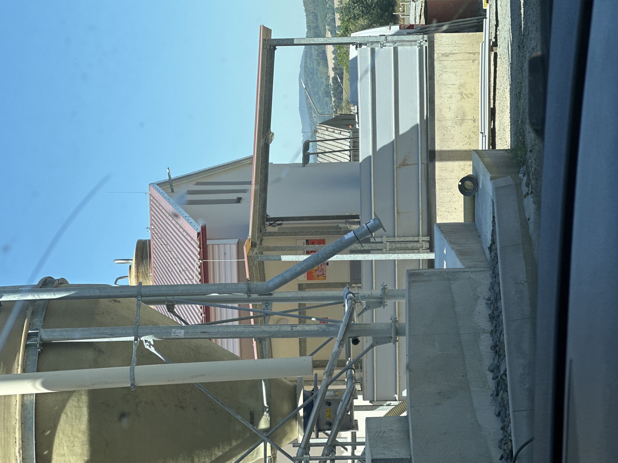 The image shows a view of industrial silos and a building, possibly a farm or agricultural facility, under a clear blue sky. In the foreground, a large, pale yellow silo is partially visible on the left, supported by metal beams. Adjacent to it, a white pipe runs vertically. A concrete structure with gravel and rocks is also present in the immediate foreground.
In the mid-ground, a white building with a reddish-brown roof is visible, featuring several vertical window-like elements. A covered walkway or extension with a metal frame and a translucent roof connects to this building. Below this, a long, light gray industrial container or structure is situated on a concrete base. A yellow corrugated pipe emerges from a concrete surface in the lower right portion of the mid-ground.
The background features a rural landscape with rolling hills covered in trees and fields. Several other structures, including pitched-roof buildings and what appear to be streetlights or poles, are scattered across the landscape. The overall impression is one of an operational agricultural or industrial site in a rural setting. The specific location in Petrovičky, Czechia, is not directly identifiable from visual cues within the image itself but is provided as context.
