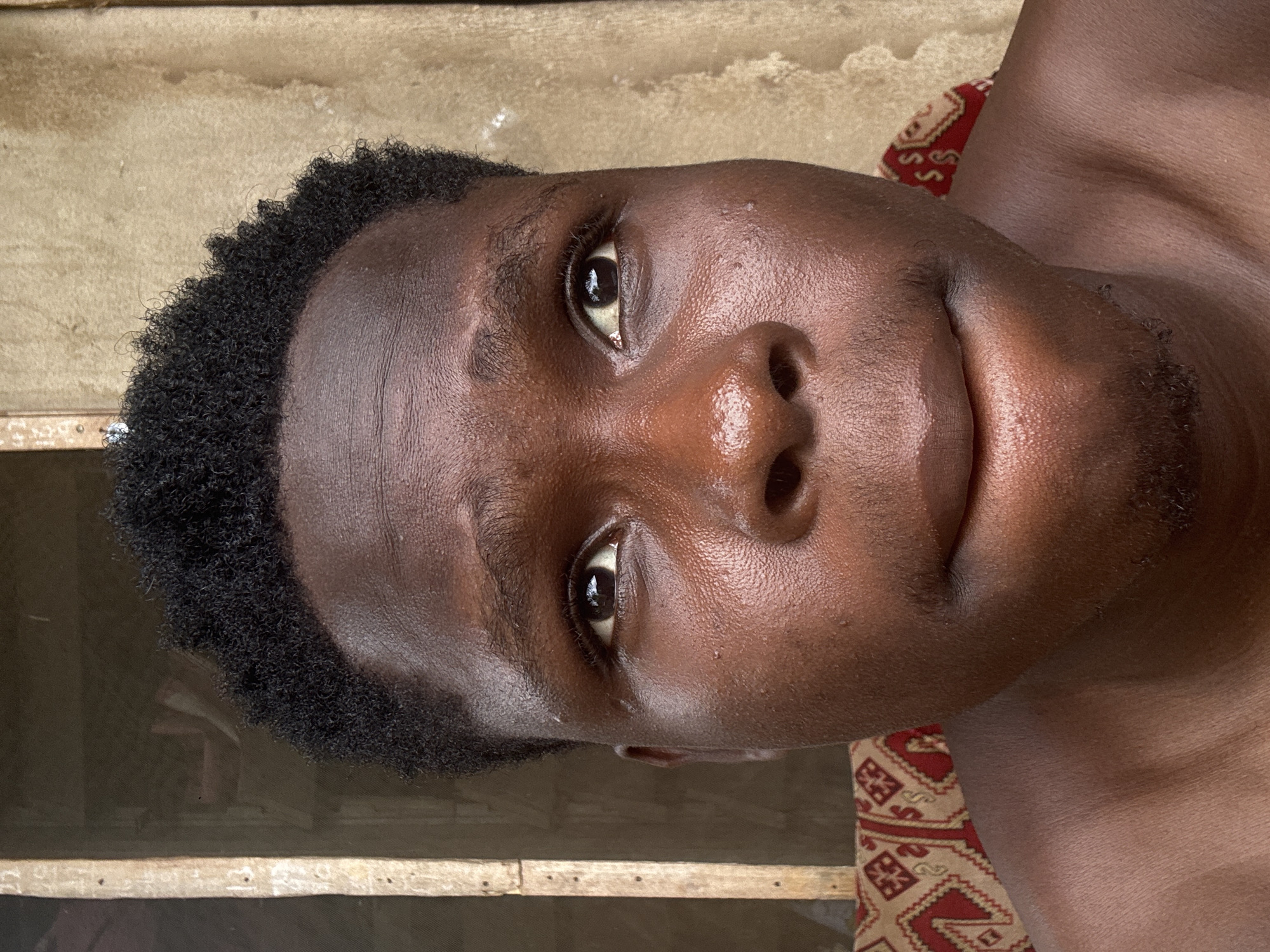 A close-up portrait of a young Black man with short, tightly curled black hair. His skin is dark brown with a slightly oily sheen. He has brown eyes. He is looking directly at the camera with a slight smile. A sparse, dark beard is visible. The background is blurred, showing a section of weathered wood and part of what appears to be a patterned fabric. There is no concrete evidence to establish the location is Aboh, Nigeria.