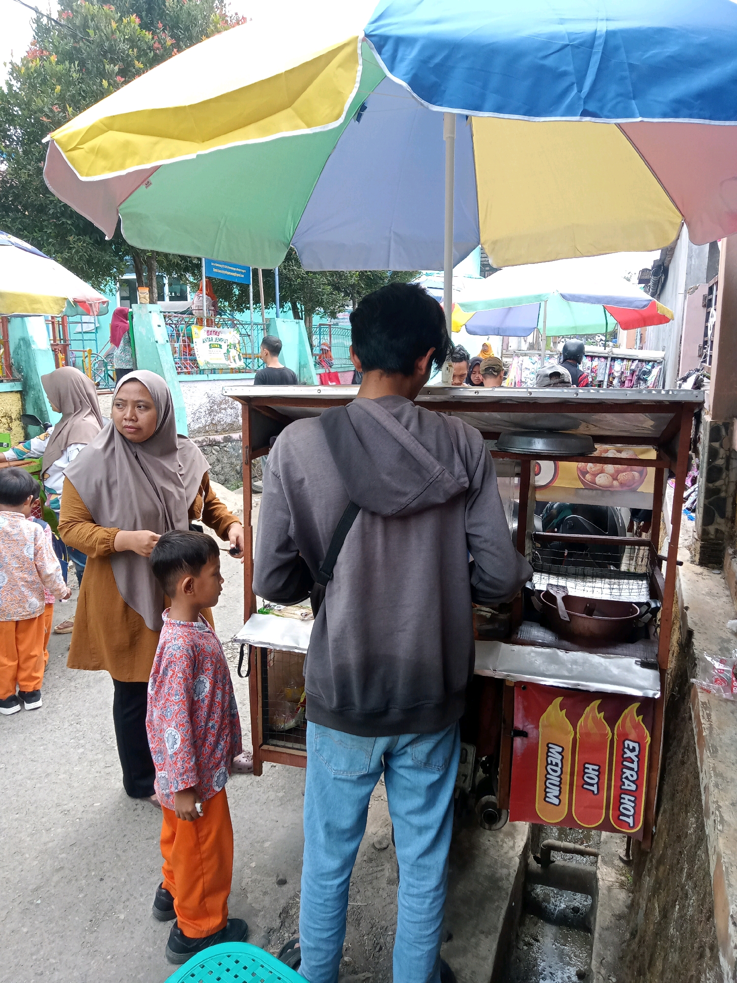 An outdoor scene in Gumiwang, Indonesia, features a food cart with a colorful umbrella providing shade. Several people are present. A woman wearing a hijab and a brown tunic stands near the cart, holding a pair of scissors. A young boy in an orange shirt and pants stands next to her. To the right, a person wearing a grey hoodie and blue jeans is facing the cart. Behind them, other individuals are visible, some under the umbrella and others further back. The food cart itself displays various items, including what appears to be a cooking apparatus and a sign with three chili pepper illustrations labeled "MEDIUM," "HOT," and "EXTRA HOT." The ground is paved, and a narrow drainage ditch runs alongside the cart. In the background, a playground structure and trees are visible, suggesting a public area.