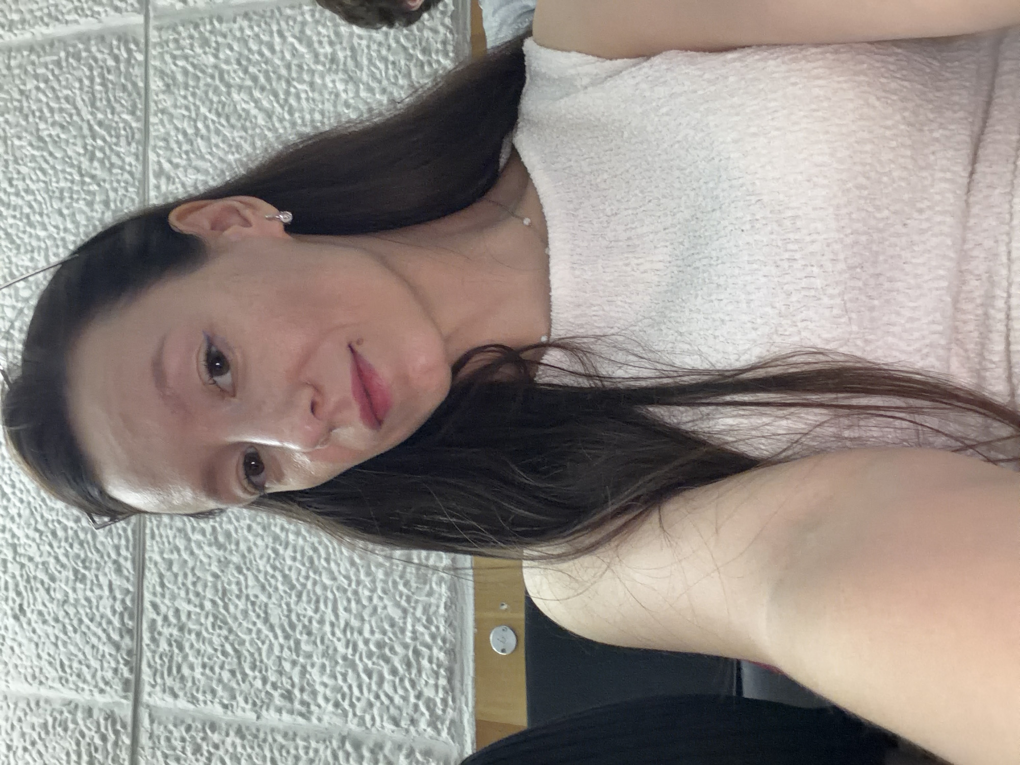 The media file is a portrait of a young woman taken from a slightly high angle, rotated counter-clockwise. She has long, dark brown hair that falls around her shoulders and a white, textured top. She is wearing subtle makeup, with pink lipstick and what appears to be eyeliner. A delicate necklace with small pearls is visible around her neck. She has a small, stud earring in her visible ear. Her expression is neutral.
The background consists of a textured, light-colored wall with a grid-like pattern, possibly cinder blocks. To the right of the woman, there is a wooden surface with what looks like a small, circular metal fixture. The lighting is soft, suggesting an indoor setting, possibly during the day. The overall mood of the image is casual and personal, like a selfie. There is no visible text in the image.
