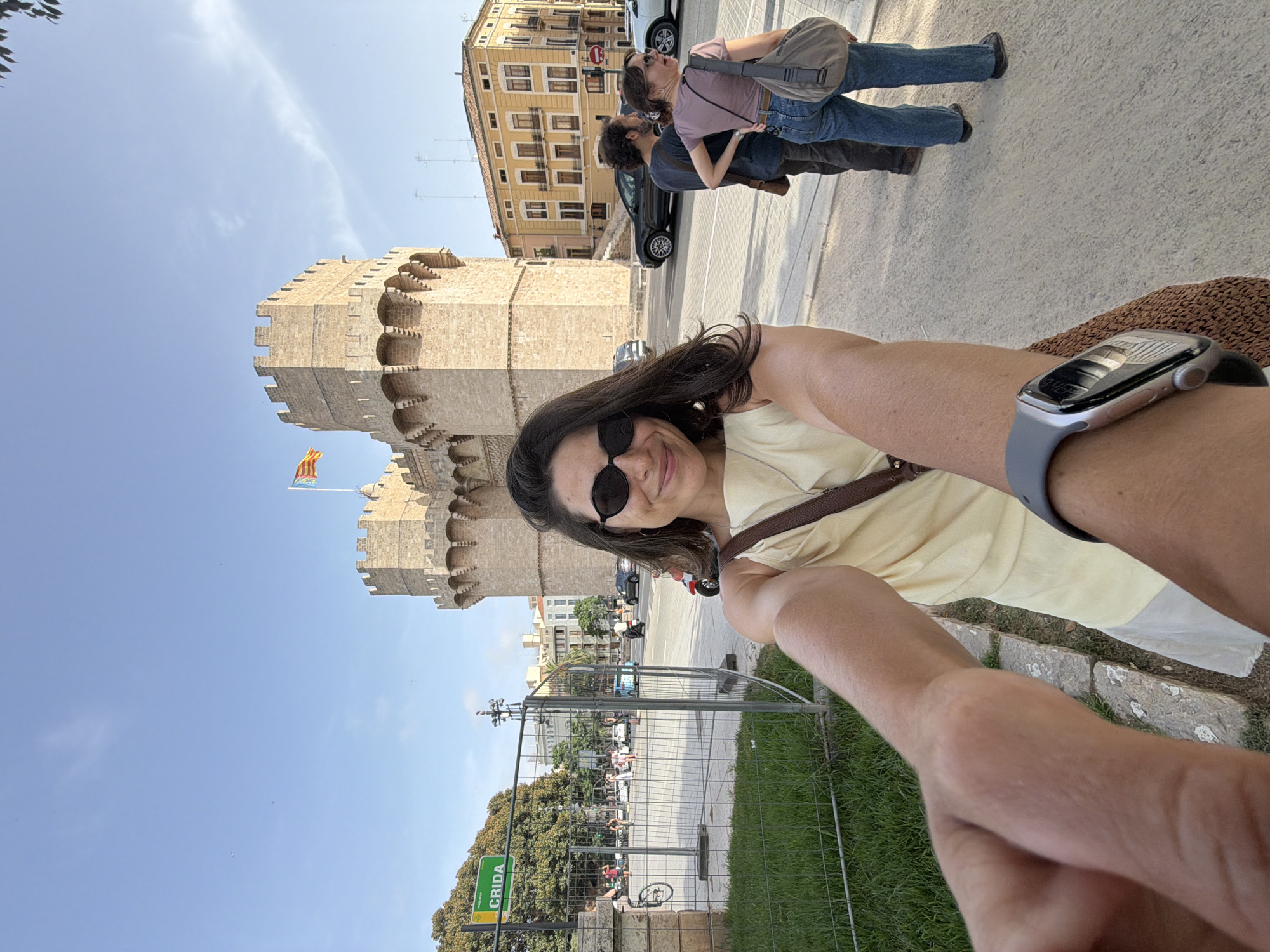 In this full-body selfie, a woman with dark hair and sunglasses smiles at the camera, holding the camera out with her right arm. She is wearing a light yellow sleeveless dress and a brown crossbody bag. Her left arm, extended towards the camera, shows a silver smartwatch on her wrist. Behind her, two people stand on a sidewalk. The man wears a dark shirt and brown bag, and the woman wears a pink t-shirt and blue jeans with a gray shoulder bag. The background features a large, ancient stone tower with battlements and a flag flying from its top, a yellow building, a street with cars and motorcycles, and a fenced-off area with trees and a sign that reads "CRIDA". The scene is set in an urban environment on a clear day with a blue sky.