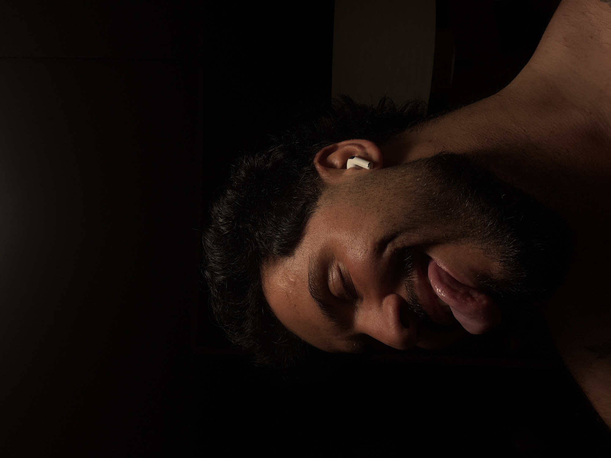 A dark image shows a person's head, tilted, with their tongue sticking out. The person has dark hair and a beard. There is a white earbud in their ear. The image is very dark, with most of the background black. It is not possible to determine the location from this image.
