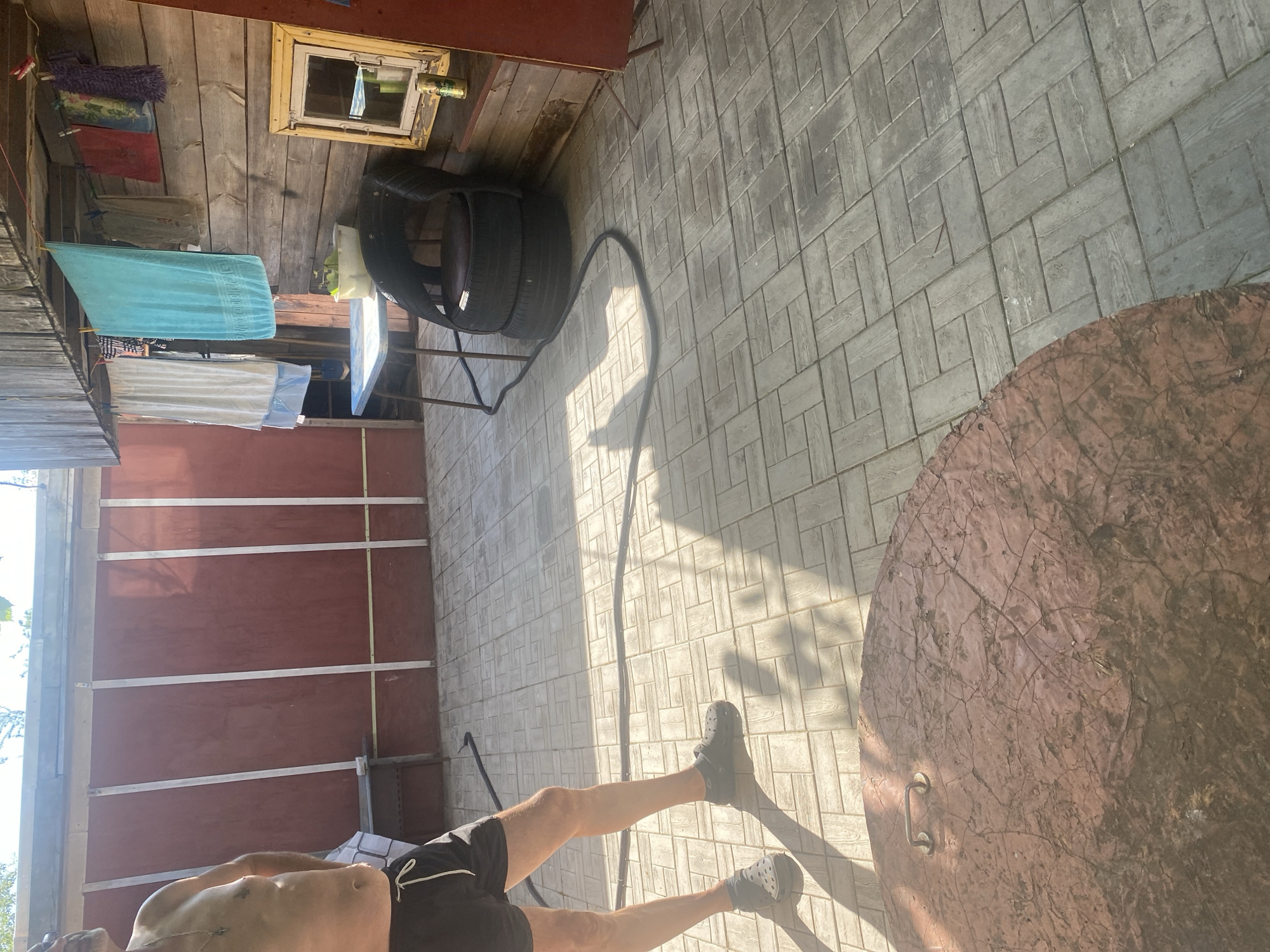 The image depicts an outdoor patio area, possibly a backyard or a dacha, in Krupki, Belarus. A partially visible man, wearing black shorts and dark clogs, stands on a tiled patio. Sunlight casts shadows across the ground. In the background, a wooden wall with hanging laundry is visible, along with a small table and a chair made from stacked tires. A circular, reddish-brown object, likely a lid or cover, is present in the foreground. The overall scene suggests a casual, rustic setting.