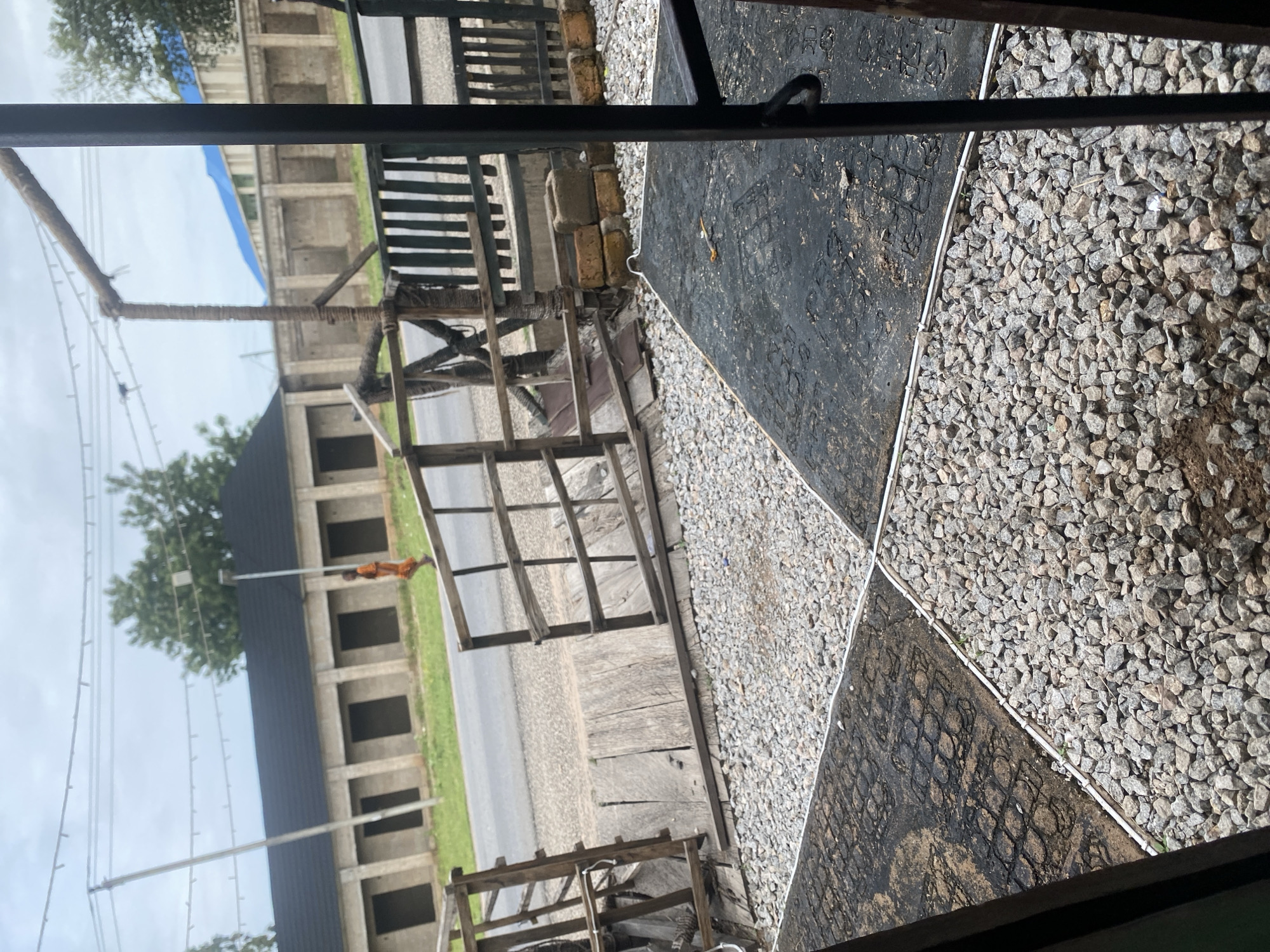 The image depicts an outdoor scene in Jalingo, Nigeria, likely during the daytime under overcast skies. In the foreground, the view is partially obstructed by a dark metal bar, suggesting the perspective is from within a structure or under an overhang. The ground is paved with a mosaic of textures: sections of dark, patterned material, possibly tar or rubber, and areas covered in small, light-colored gravel. A white pipe runs along the edge of these paved sections.
Beyond the immediate foreground, there is a wooden structure, perhaps a gazebo or shelter, with exposed beams and a slatted roof. Next to this, a gravel path continues. In the middle ground, a paved road is visible, bordered by grassy verges. Across the road stands a building under construction, with a dark roof and multiple open, rectangular window or door openings. Trees are visible behind the building, and the sky is a muted gray, indicating cloudy weather.
The scene appears to be in a semi-urban or developing area, given the unfinished building and the mix of paved and natural ground cover. There are no people clearly visible in the image, and no discernible text. The overall impression is one of a functional, perhaps utilitarian, outdoor space.